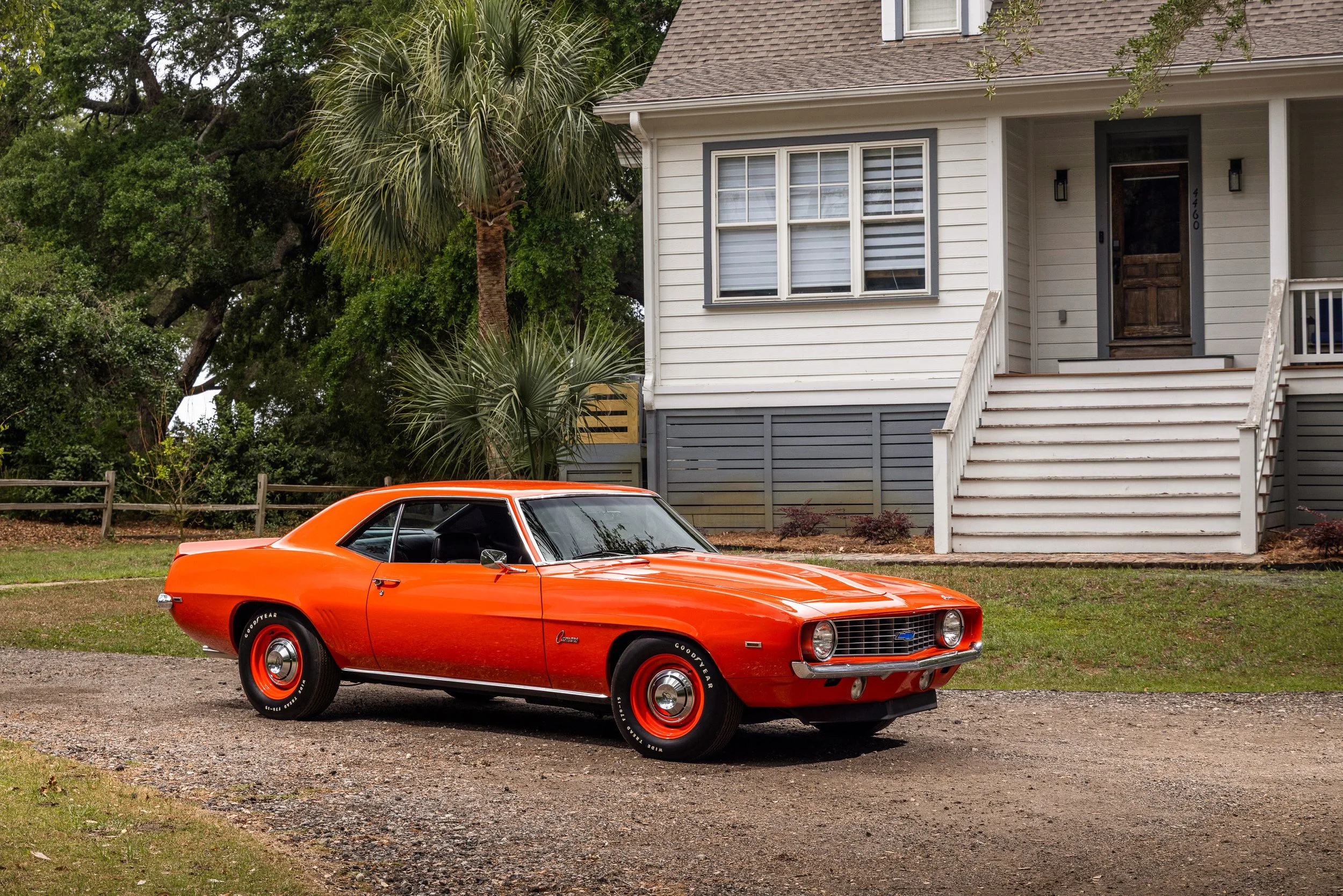 A bright orange vintage Chevrolet Camaro parked in front of a white house with stairs leading up to the front door and lush green trees in the background.