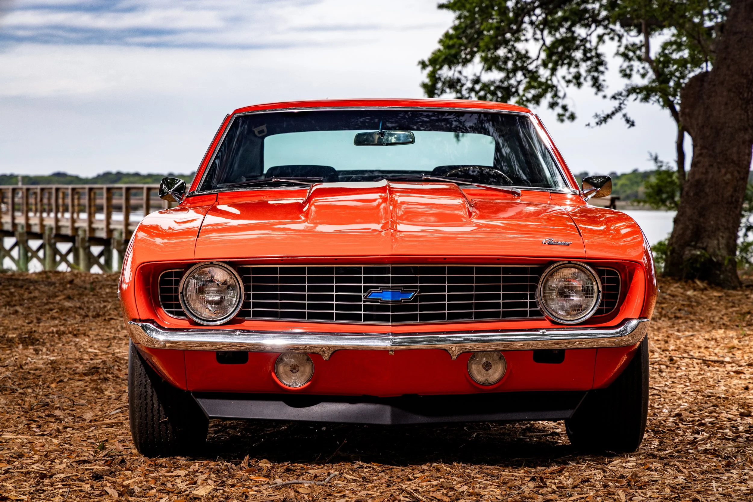 Front view of a red classic Chevrolet Camaro car parked on a dirt ground near trees, with a lake and bridge in the background.