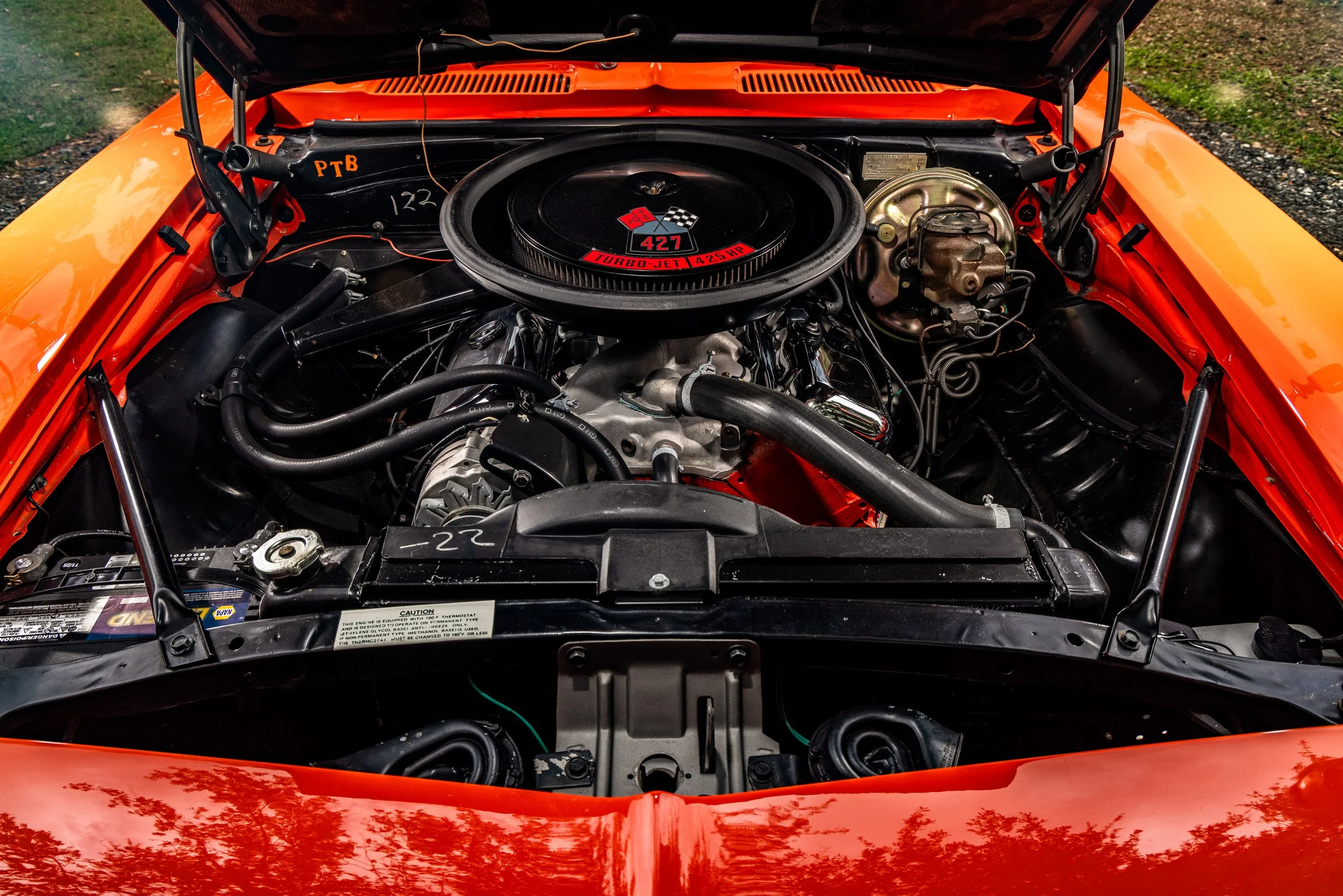 Close-up view of a vintage orange car's engine compartment with a black air cleaner cover labeled '427 Turbo-Jet 425 HP' and various engine components visible.