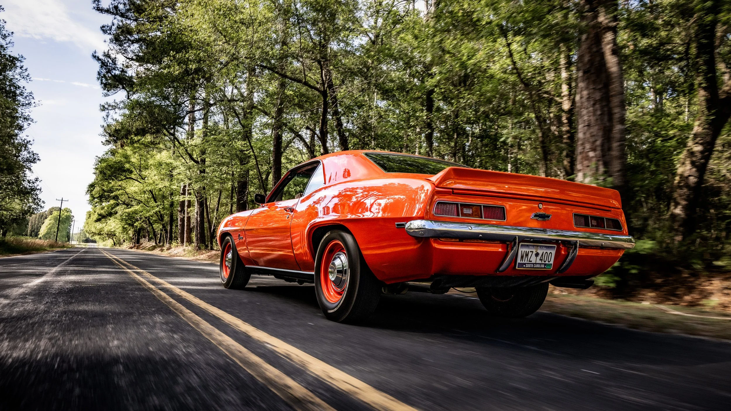 A red vintage Chevrolet car driving down a two-lane road through a forested area with green trees on both sides.