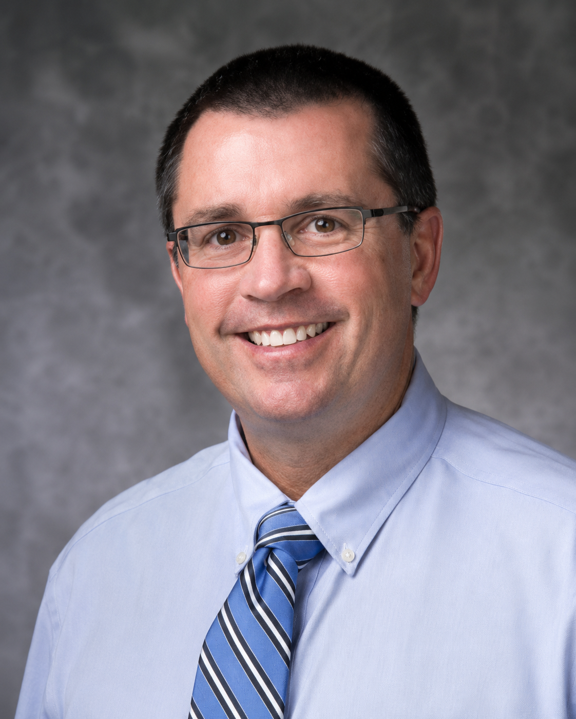 A professional headshot of a man with short dark hair, glasses, wearing a light blue dress shirt and a striped blue tie, smiling against a gray background.