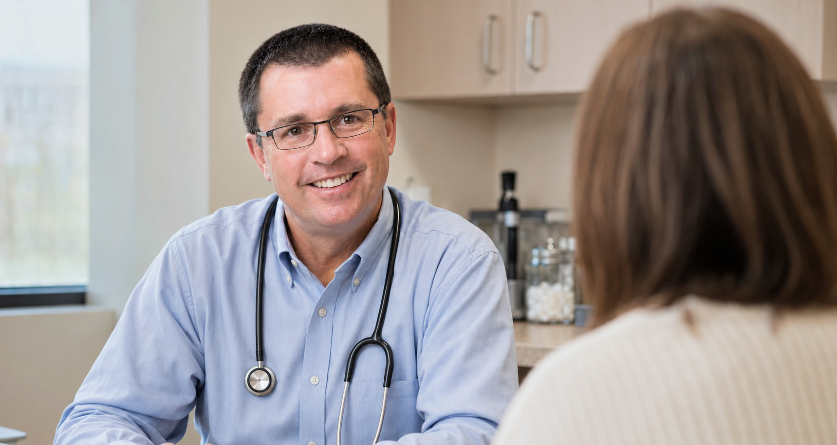 A male doctor with glasses, wearing a blue shirt and stethoscope around his neck, smiling and talking to a woman with brown hair in a beige sweater, in a medical office setting.