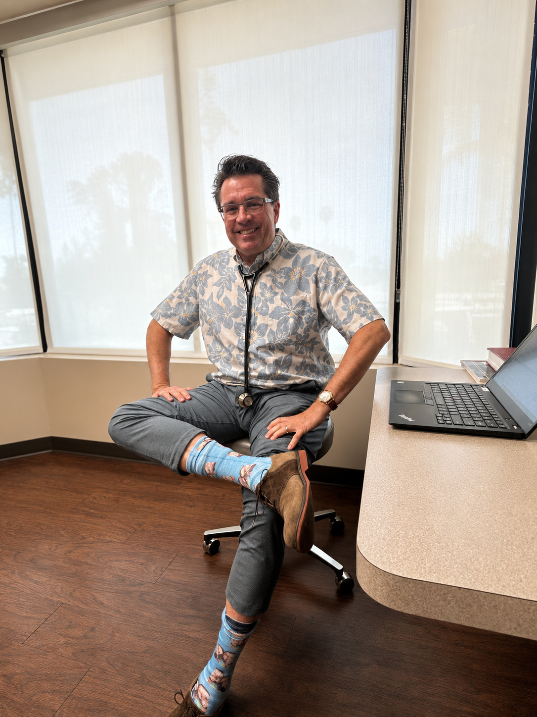 A man dressed like a doctor, wearing a stethoscope, colorful patterned socks, and casual clothes, sitting in a medical office with a laptop and large windows in the background.
