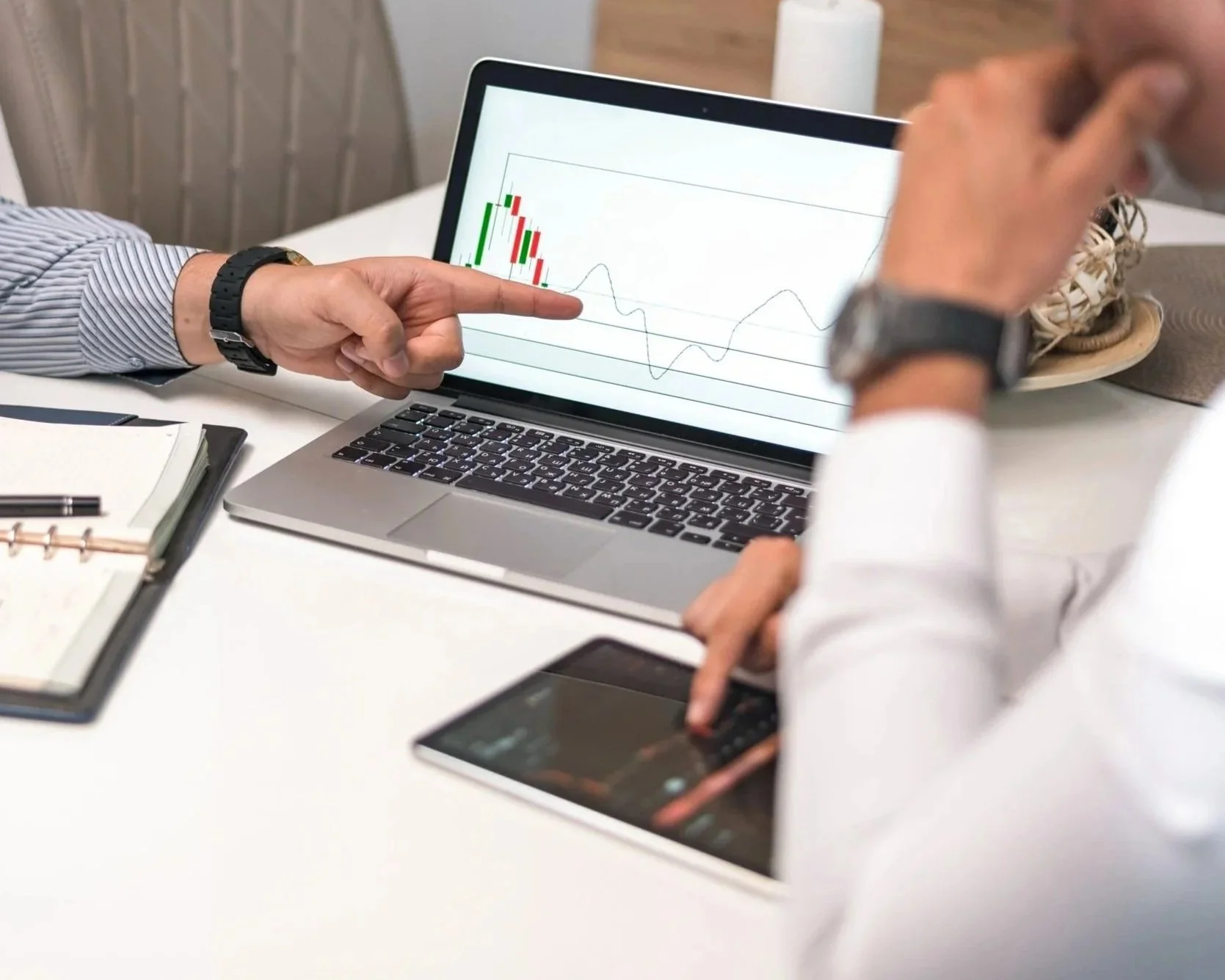Two people looking at a laptop screen displaying a financial stock chart with candlesticks and a line graph, sitting at a white desk with notebooks, a smartphone, and a cup.