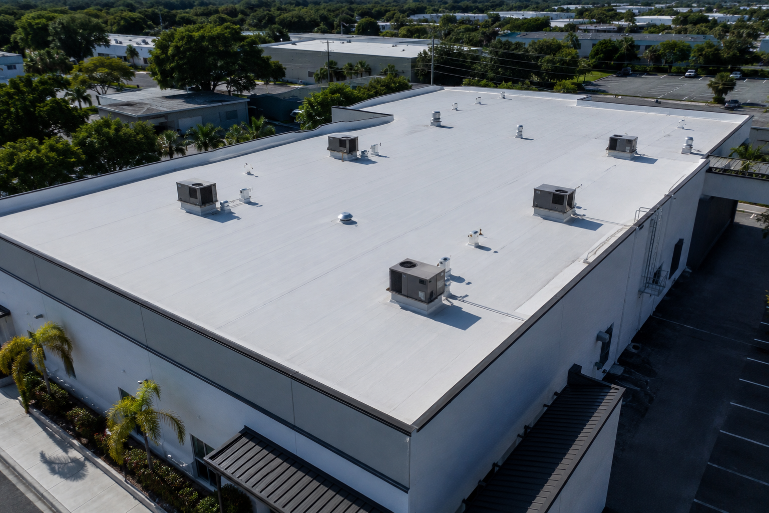 A large commercial building with a flat white roof, equipped with multiple air conditioning units and ventilation systems, surrounded by greenery and parking lot.
