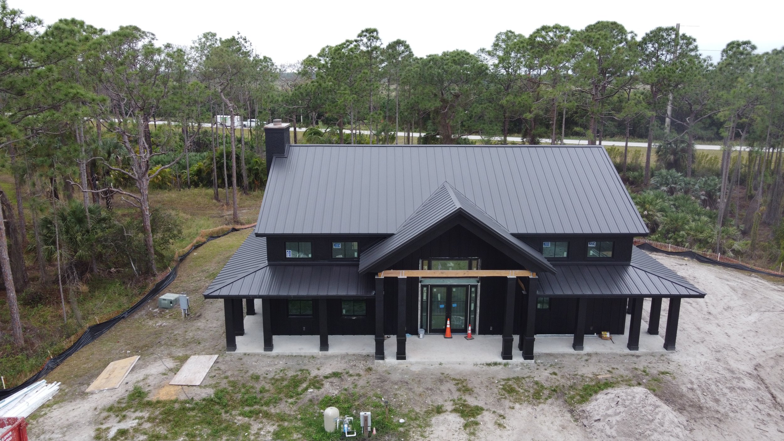 A house under construction with a black metal roof surrounded by trees and construction materials nearby.