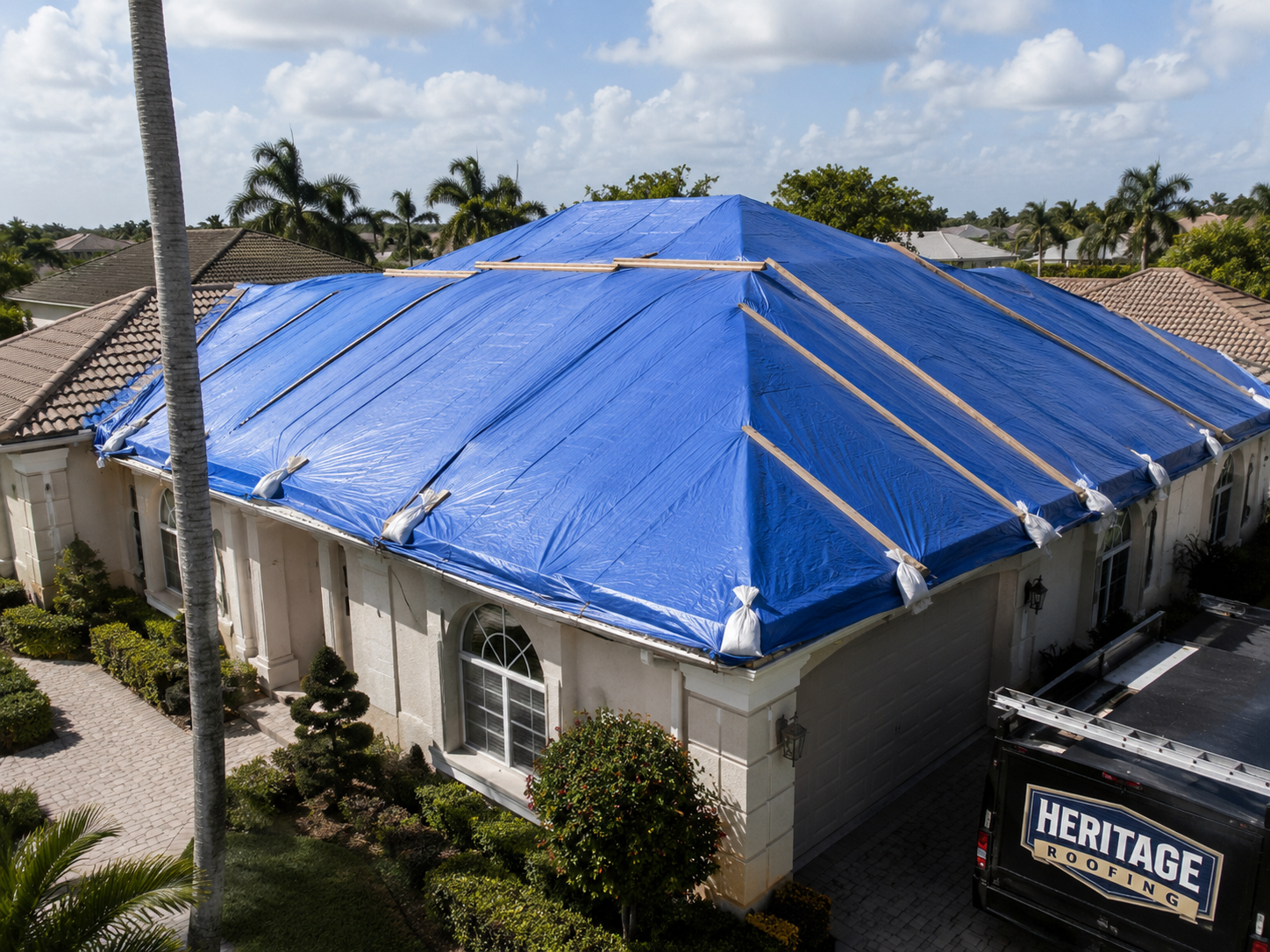 A house roof covered with a blue tarp held down by sandbags and wooden planks, likely for weather protection or repair.