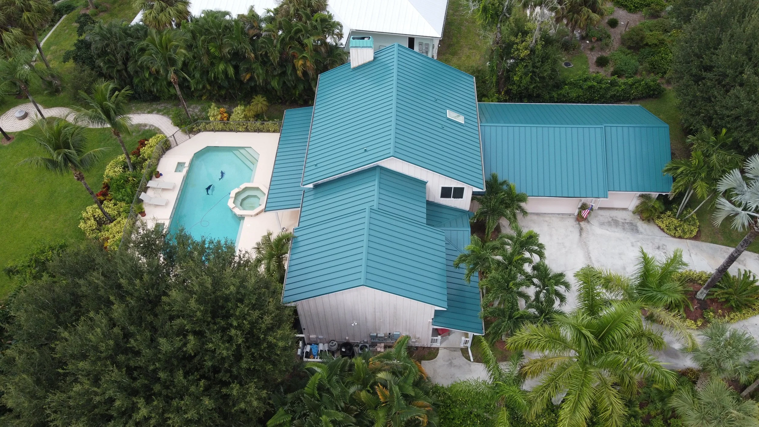 Aerial view of a house with a blue metal roof, surrounded by lush green trees and a backyard with a swimming pool and a hot tub.