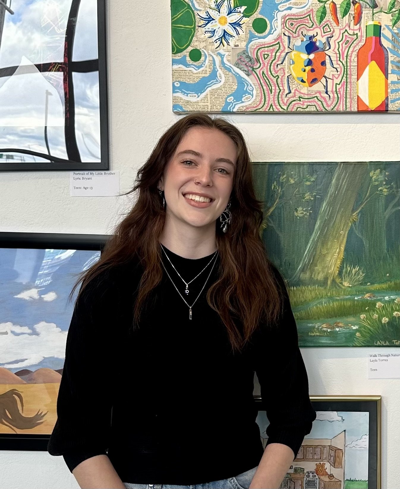 Young woman with long brown hair, wearing a black top and layered necklaces, smiling at art exhibit with colorful paintings in the background.