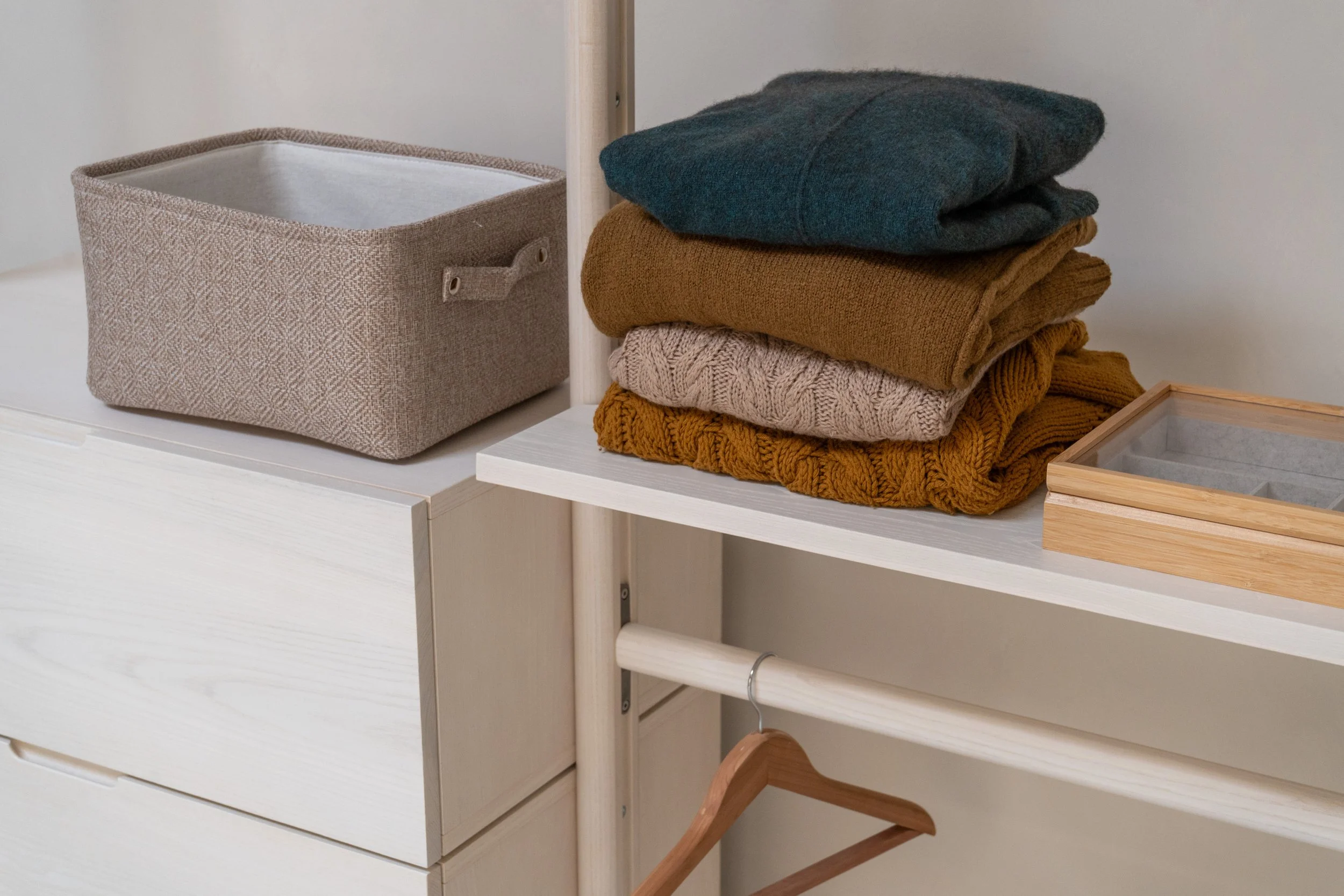 A white furniture unit with a fabric storage box on top, a stack of folded sweaters, a wooden tray, and a wooden hanger underneath.