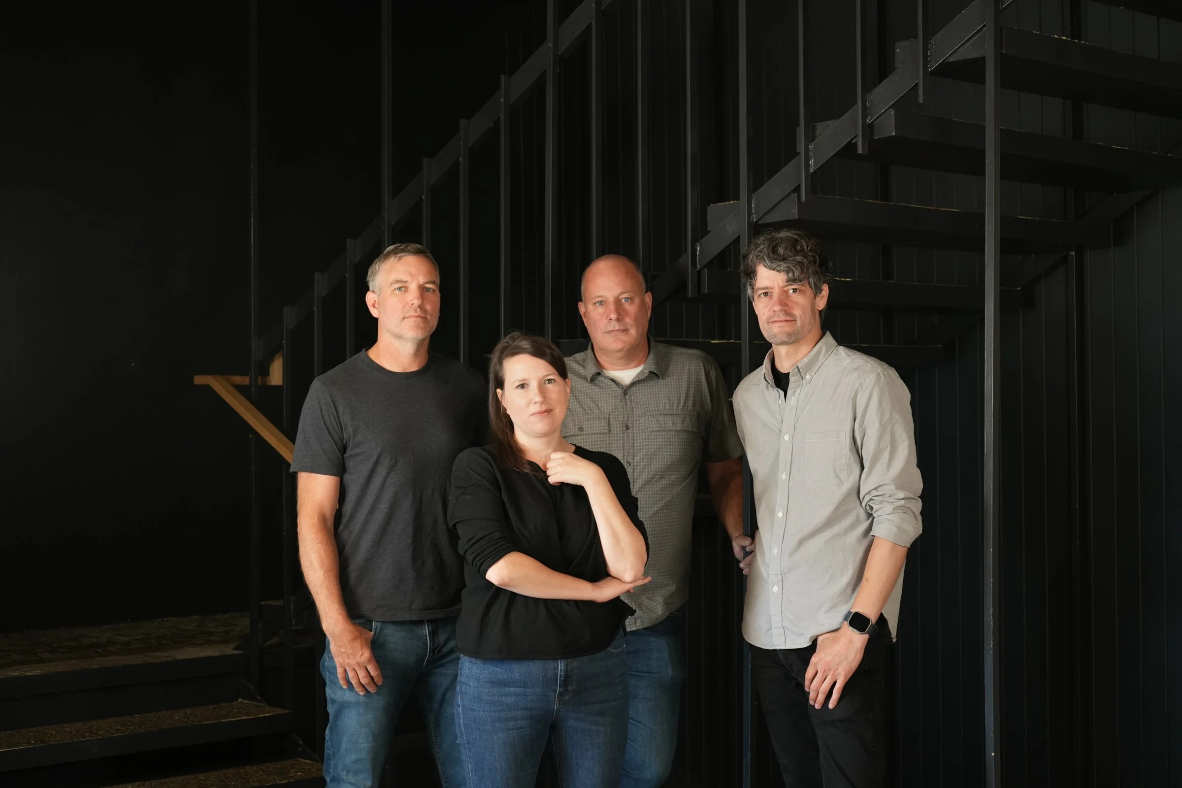 Four adults standing in front of a black staircase with black walls and railings. The group includes two men with short hair, one woman with medium-length dark hair, and a man with curly gray hair. They are dressed casually.