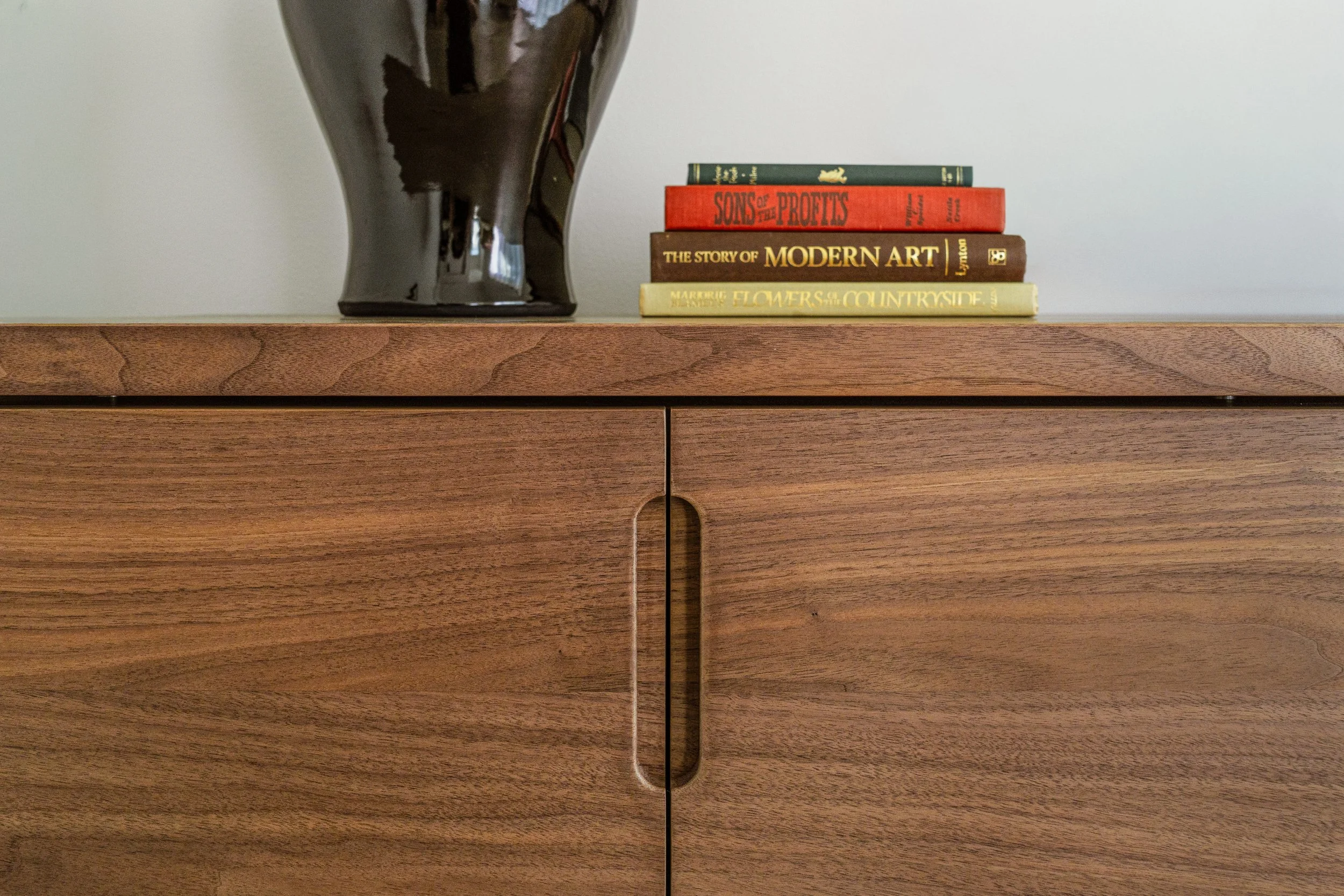 A wooden cabinet with a variety of books stacked on top, next to a black vase.