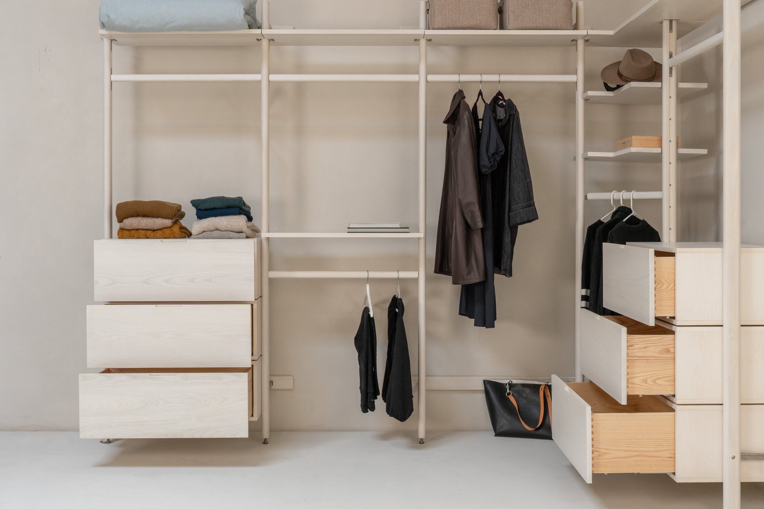 Modern, minimalist white wardrobe with various clothes, hats, and storage boxes. Folded sweaters on a drawer, hanging coats, and shoulder bags visible.