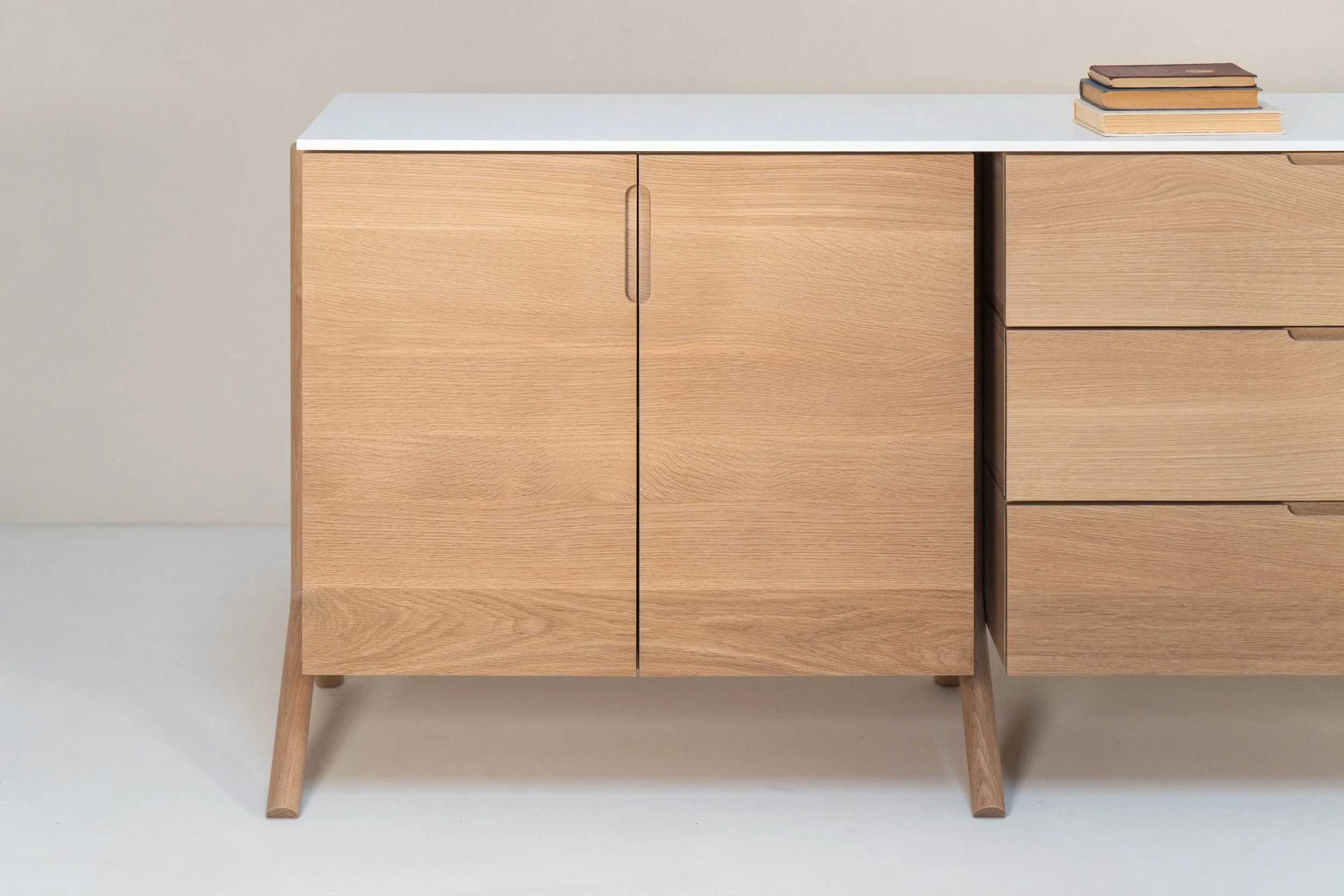 A light wood sideboard with a white top surface, four short angled legs, with three drawers on the right and a cabinet door on the left. There are four stacked books on top of the sideboard.