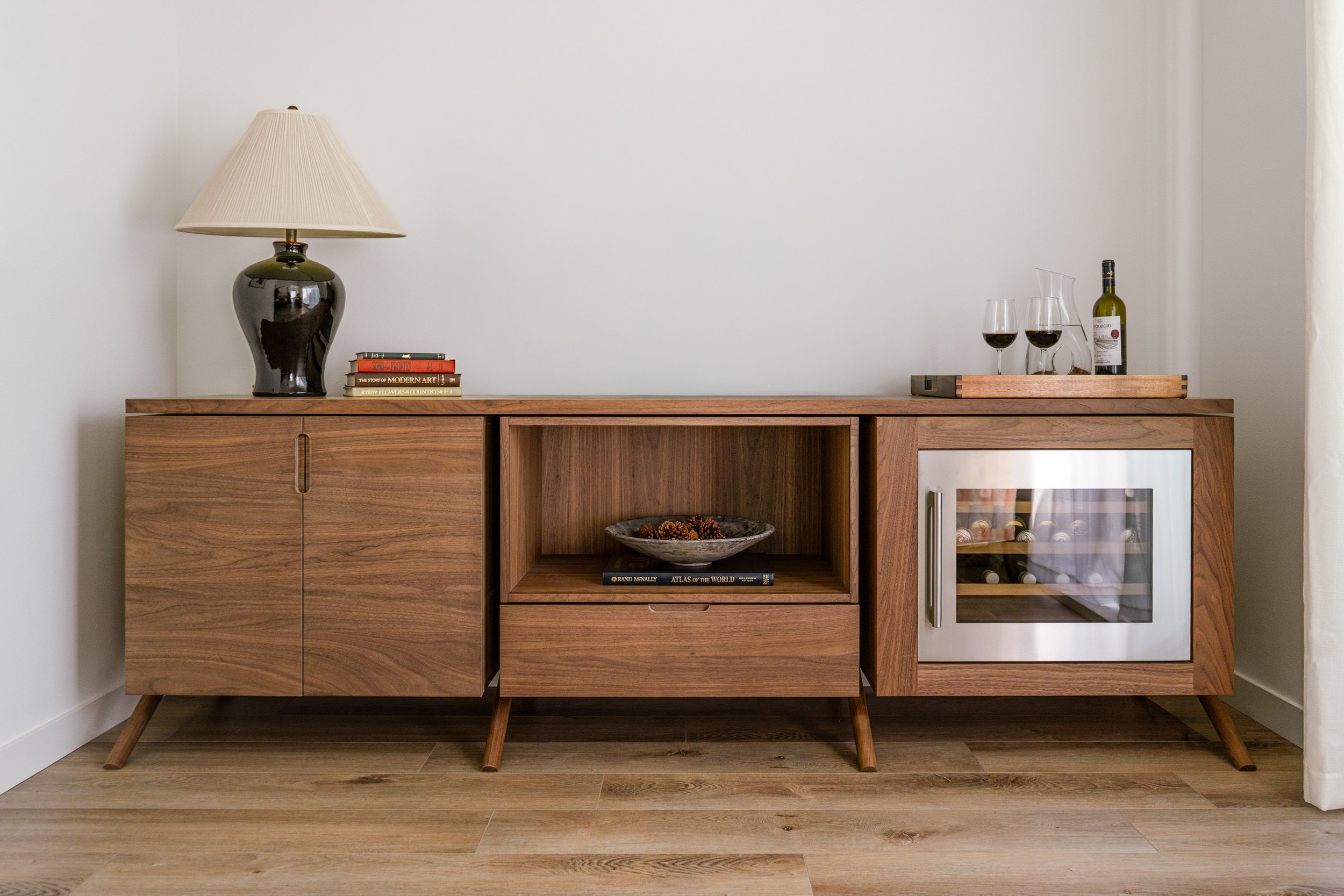 A mid-century modern wooden sideboard with tapered legs, placed against a plain white wall. On the left side of the sideboard, there is a black ceramic table lamp with a beige lampshade. Next to the lamp, there is a stack of books with titles including "Modern Art" and "Flowers of the Countryside." On the right side of the sideboard, there is a wooden tray holding two glasses of red wine, a wine bottle, and a glass decanter. Below the tray, in the open shelf, there is a decorative bowl with pinecone decor and a book titled "Atlas of the World." The floor is made of light-colored wooden planks.