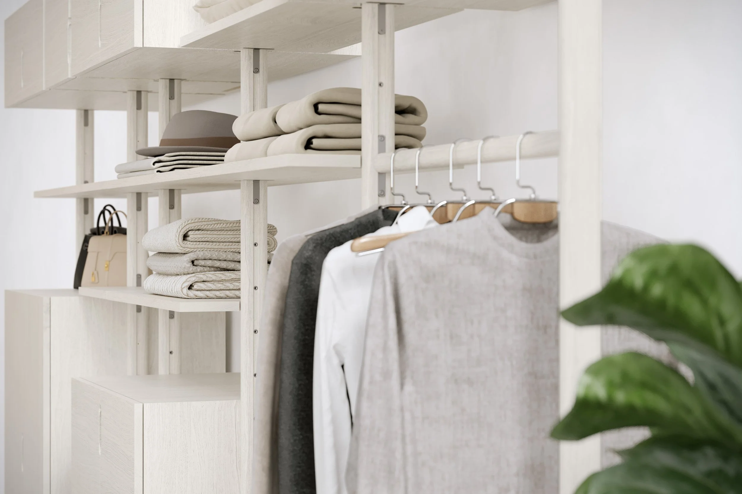 A white wooden shelf with folded beige and gray clothing, a hat, and a beige handbag, with hanging clothes below and a green plant in the foreground.