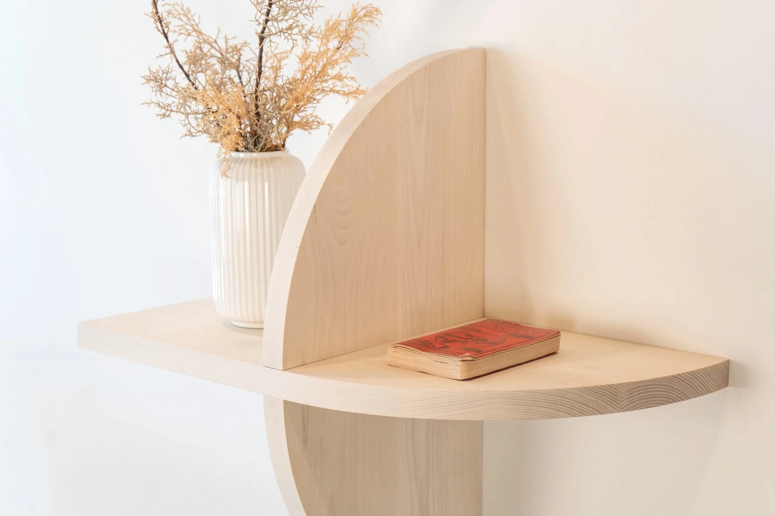 A light-colored wooden shelf with a curved edge, holding a white striped ceramic vase with dried branches, and a small stack of red and black patterned paper or cards.