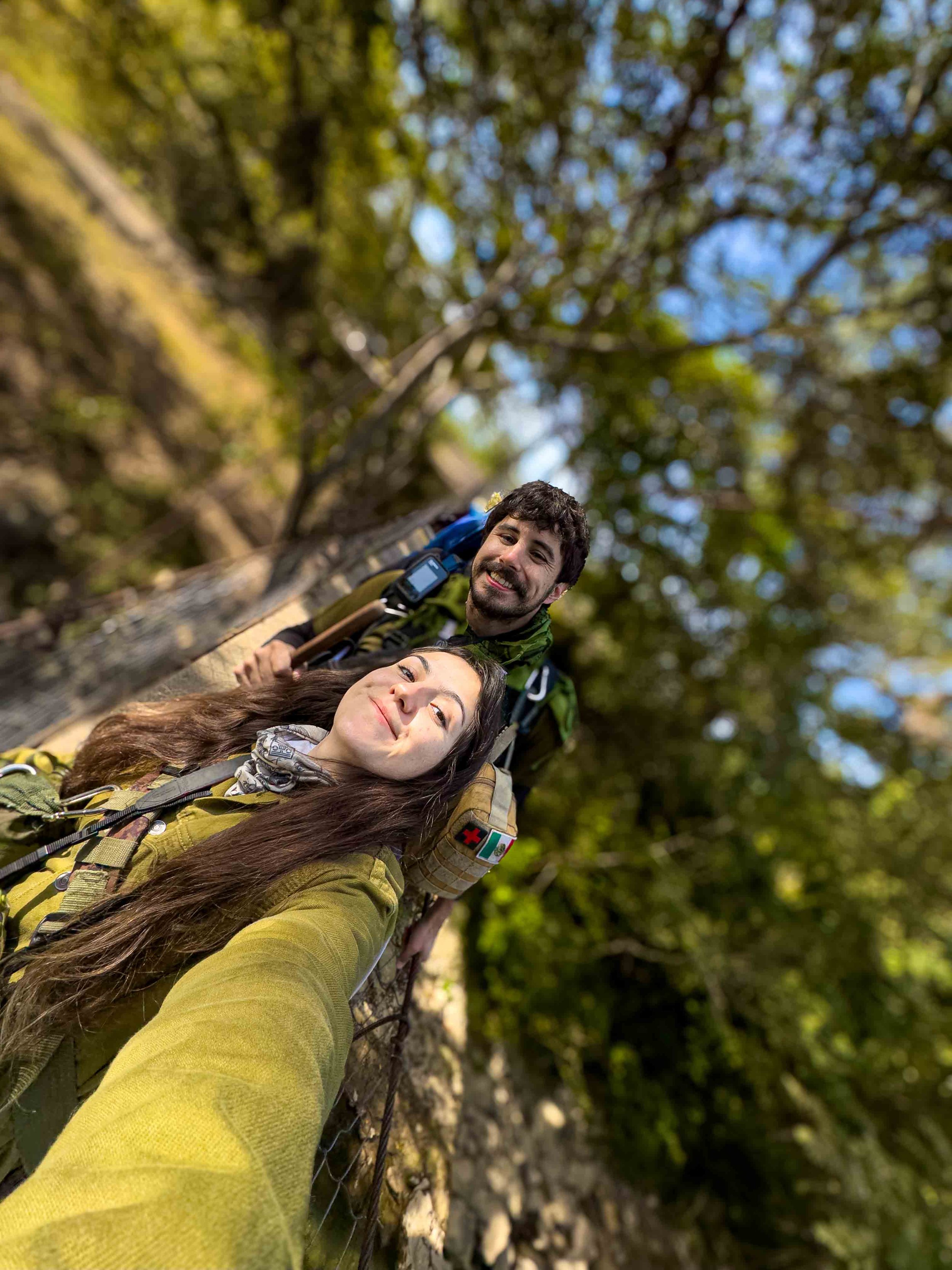 Guías de excursionismo en México, dos personas, un hombre y una mujer, tomando una selfie con un fondo de árboles y cielo azul. Ambos llevan equipo de senderismo estan en la naturaleza.