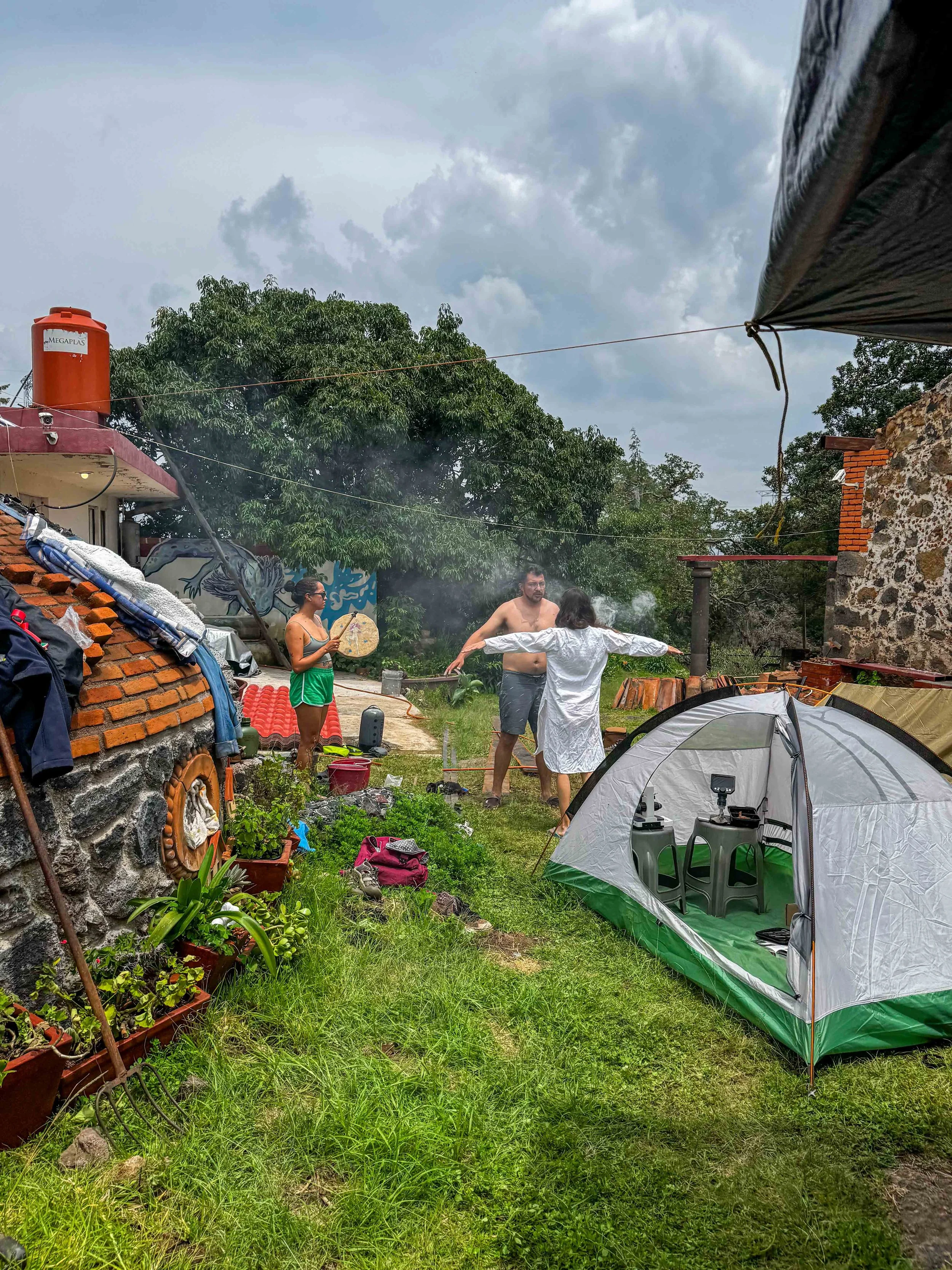 Escena al aire libre con personas en un jardín, tiendas de campaña, una estructura de piedra y ladrillos, plantas, árboles y cielo nublado. Temazcal y ciencia en rancho ajolotequio en almonte hike