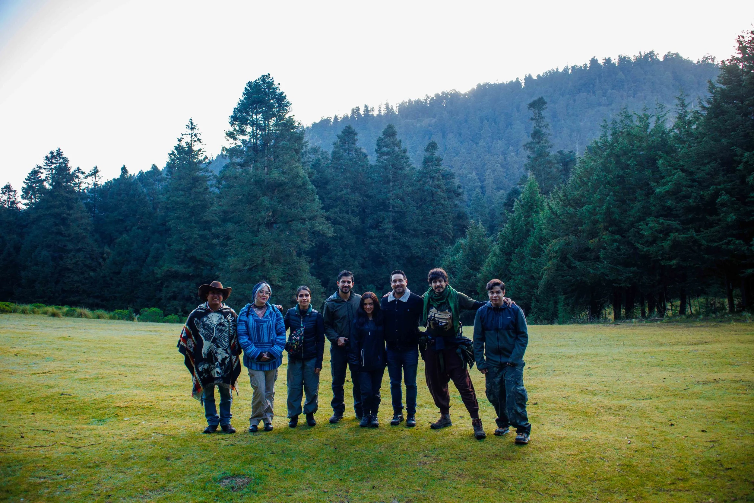 Grupo de personas de diferentes edades y géneros posando en un campo abierto con árboles y montañas de fondo, durante el día.