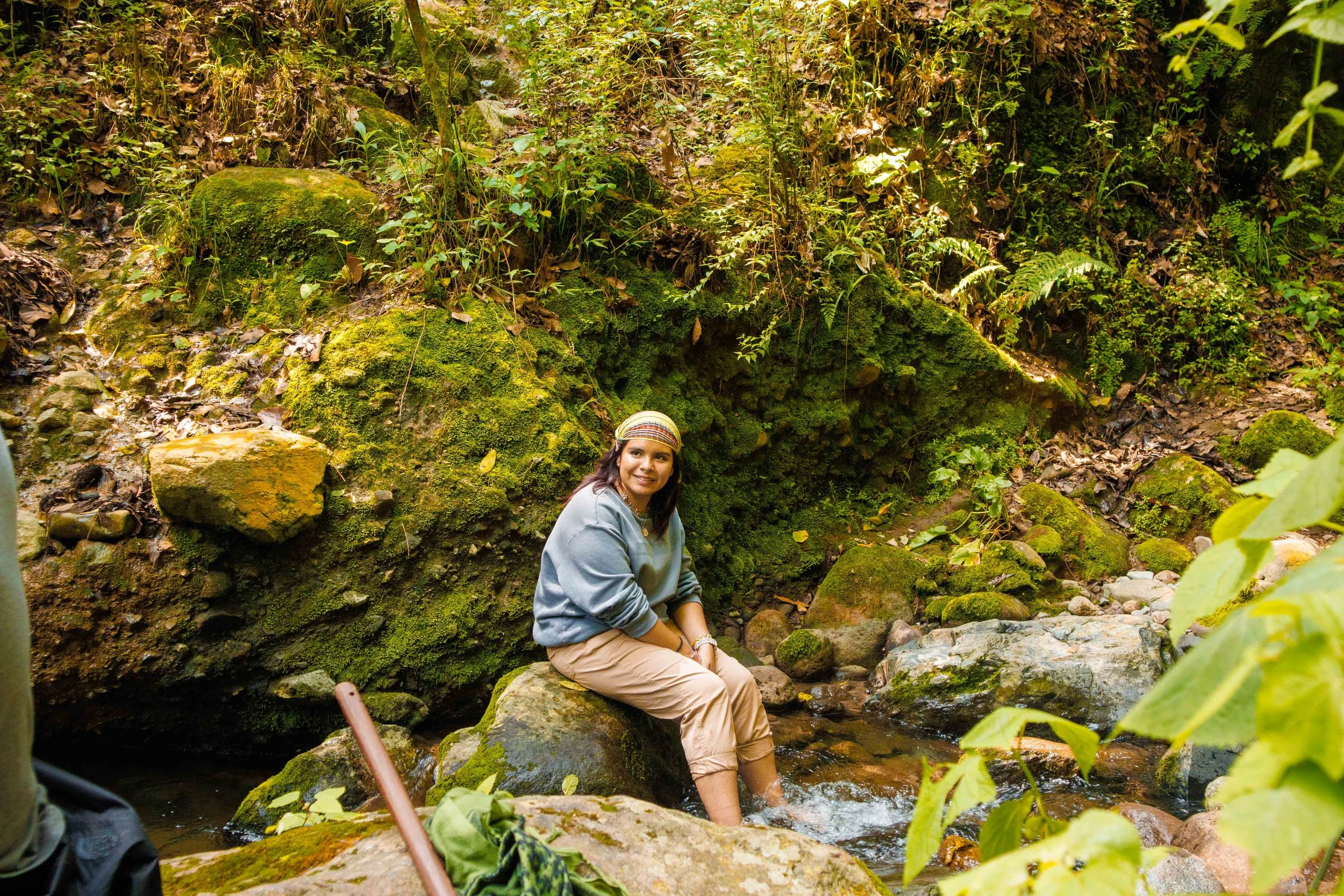 Una mujer con un gorro de colores sentada en rocas en un río de un bosque tropical rodeada de vegetación verde