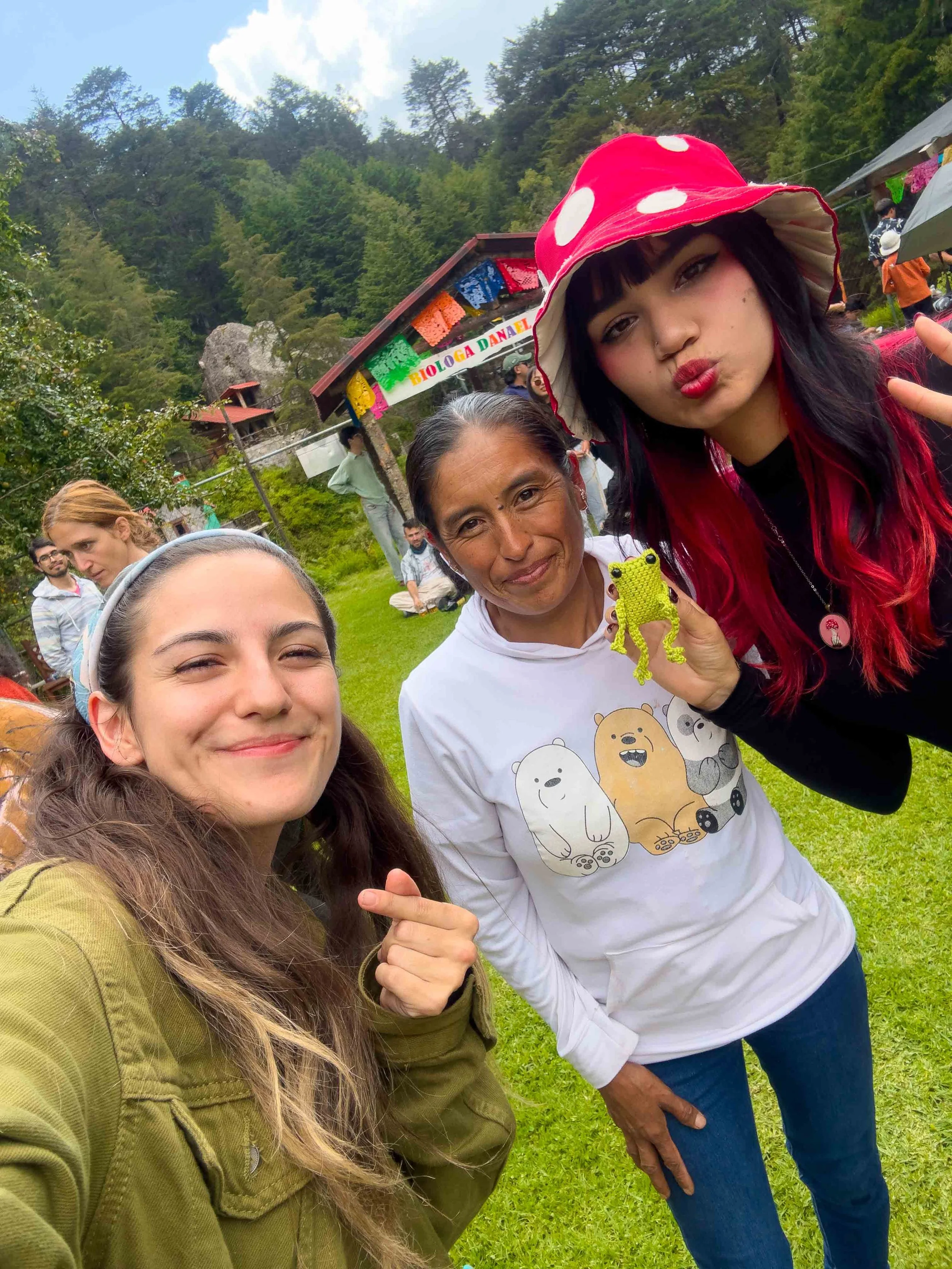 Grupo de personas en un entorno natural, algunas sonriendo y haciendo gestos, una mujer con sombrero de mujer de color rojo, otra mujer con camiseta con osos y una figura de tela verde en forma de rana, en un área verde con árboles y estructuras rura