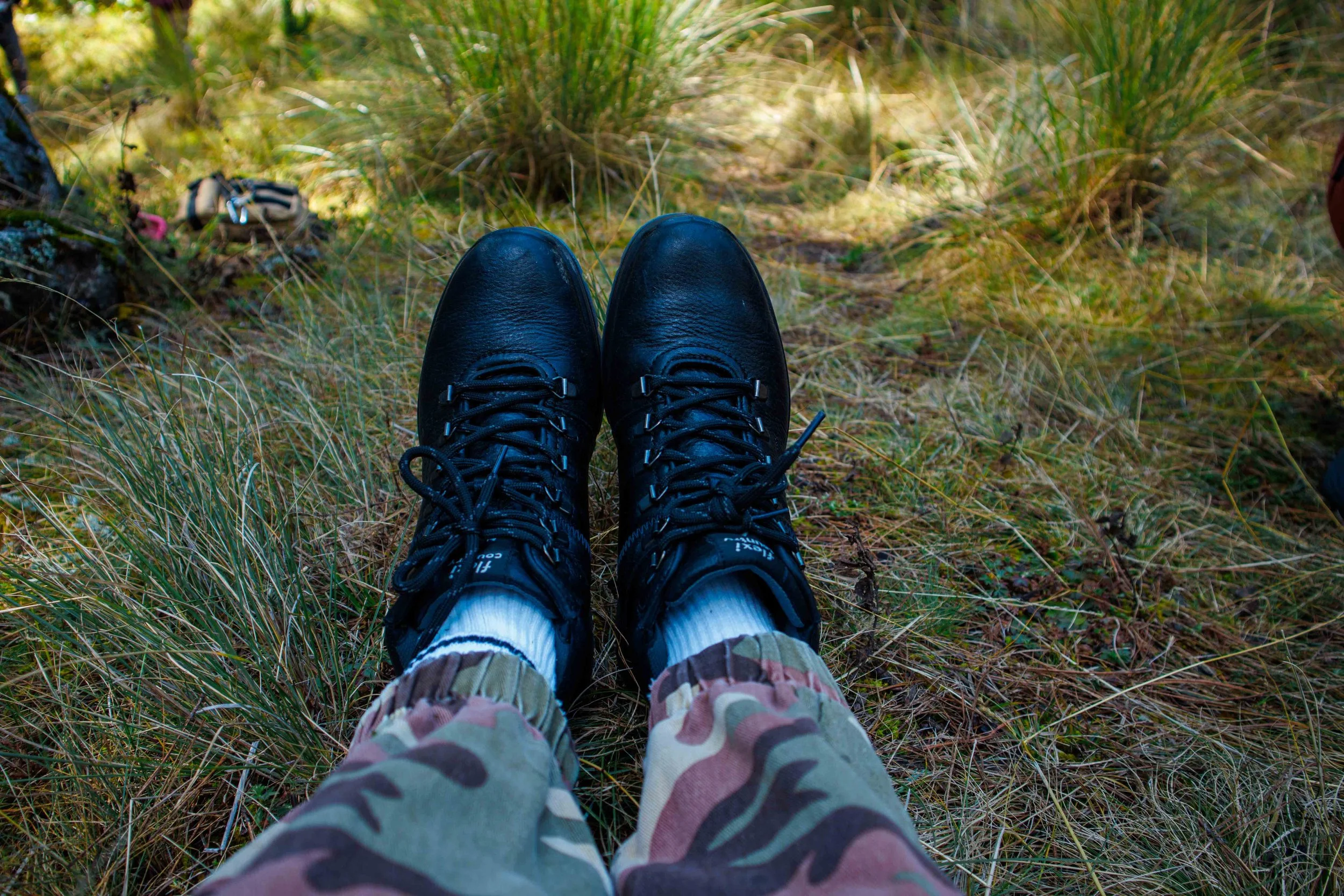 Zapatos negros de senderismo vistos desde los pies, con pantalones de camuflaje, en un sendero de tierra cubierto de pasto y vegetación, en un entorno natural al aire libre.