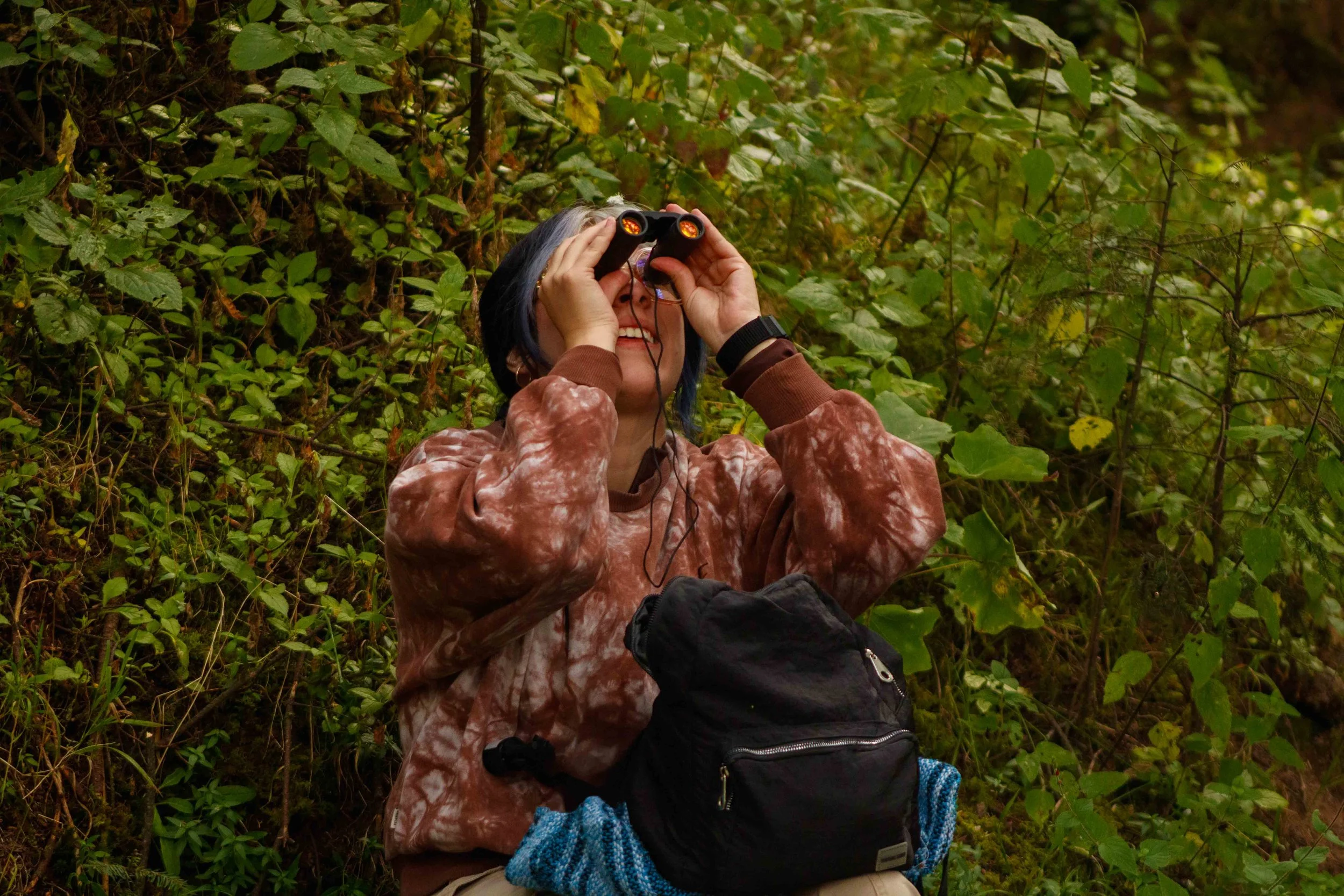 Una persona con cabello gris y oscuro usando binoculares en medio de la vegetación, con mochila y ropa de manga larga de color marrón. Está en una zona de bosque o arbustos.
