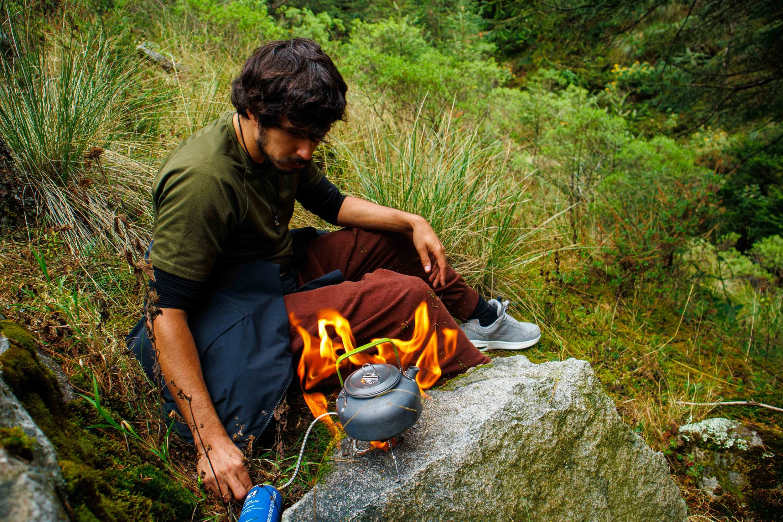 Persona sentada junto a una fogata en un entorno de bosque, con utensilios de campamento, rodeada de vegetación verde.