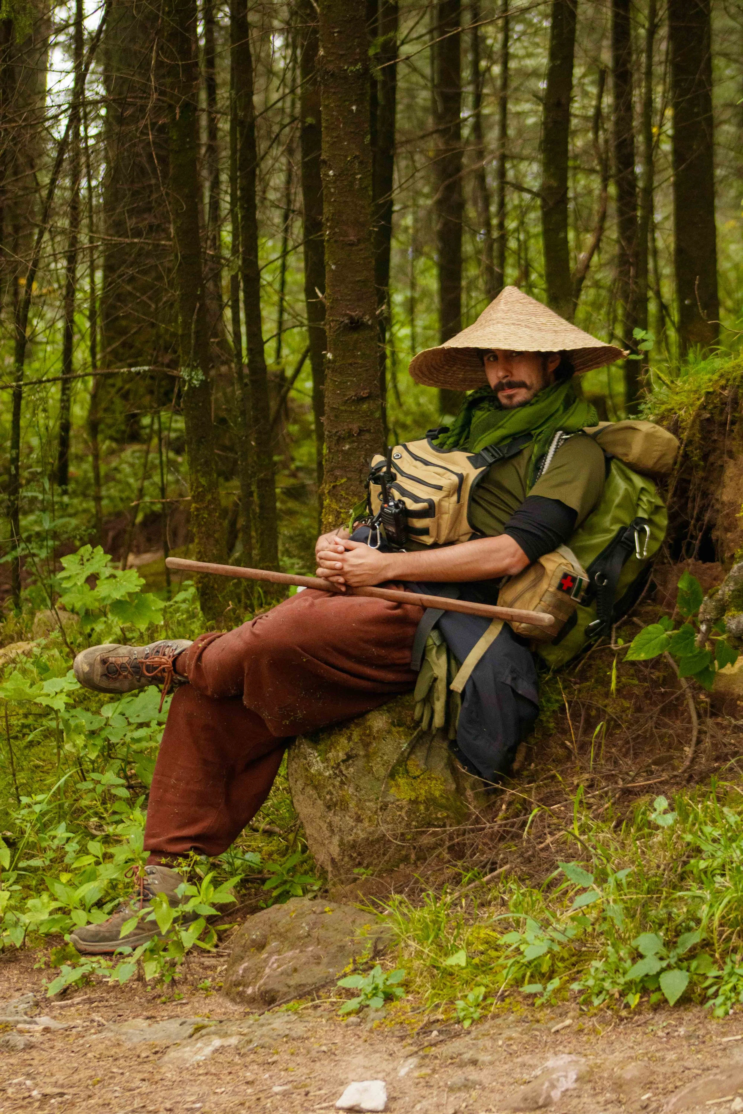 Hombre sentado en un árbol en un bosque, lleva sombrero de paja, mochila y pantalones marrones, con un bastón y ropa de montaña.