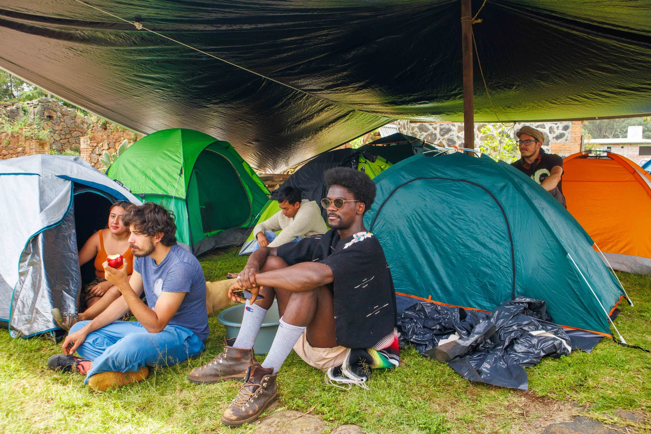 Grupo de jóvenes acampando bajo una lona, con muchas tiendas de campaña coloridas alrededor, en un entorno natural con plantas y rocas.