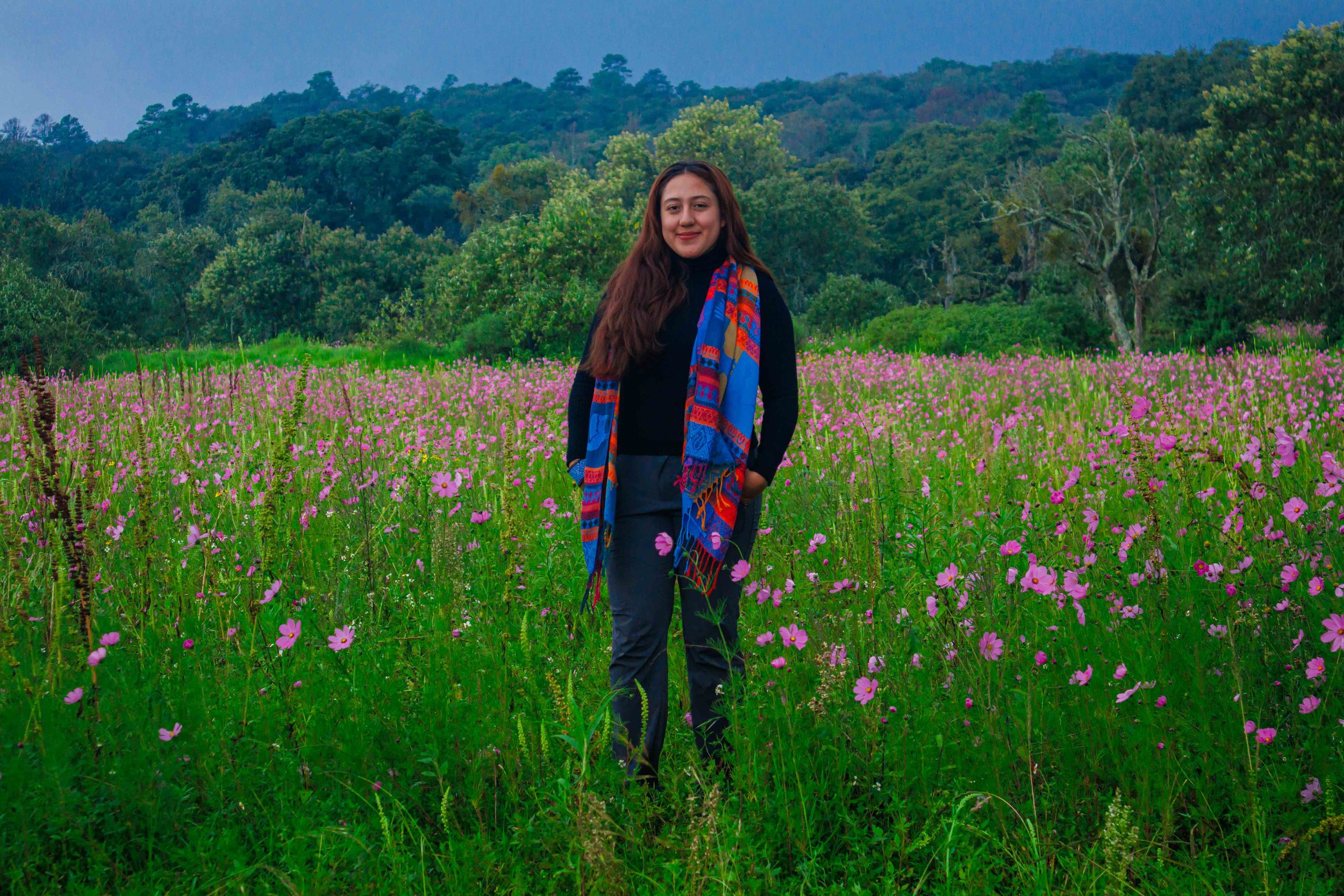 Mujer con bufanda colorida en campo de flores rosas, con árboles y colinas en el fondo, cielo nublado.