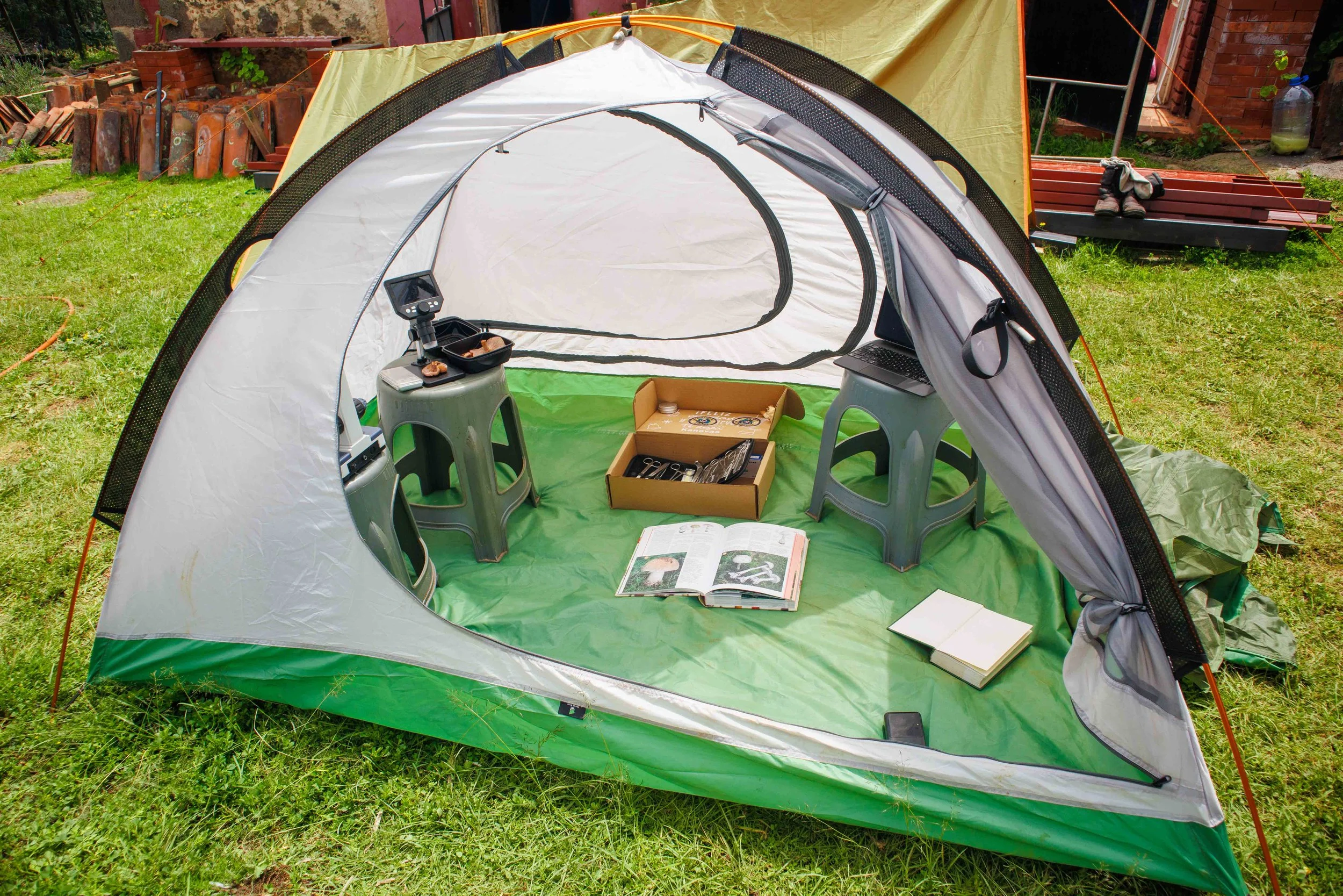 Puesto de ciencia: Una tienda de campaña en un jardín con sillas, una caja con utensilios, libros abiertos y otros objetos dispersos en el interior