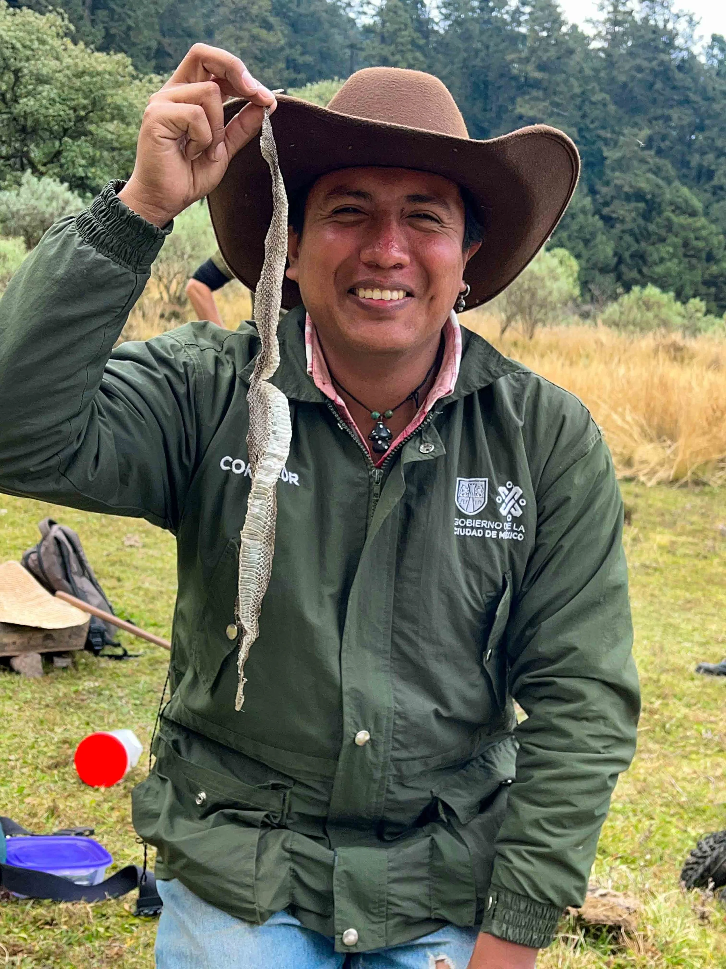 Hombre sonriendo, lleva sombrero grande, chaqueta verde con logotipo del Gobierno de la Ciudad de México, en un entorno natural con árboles y pasto, sostiene un serpiente con su mano.