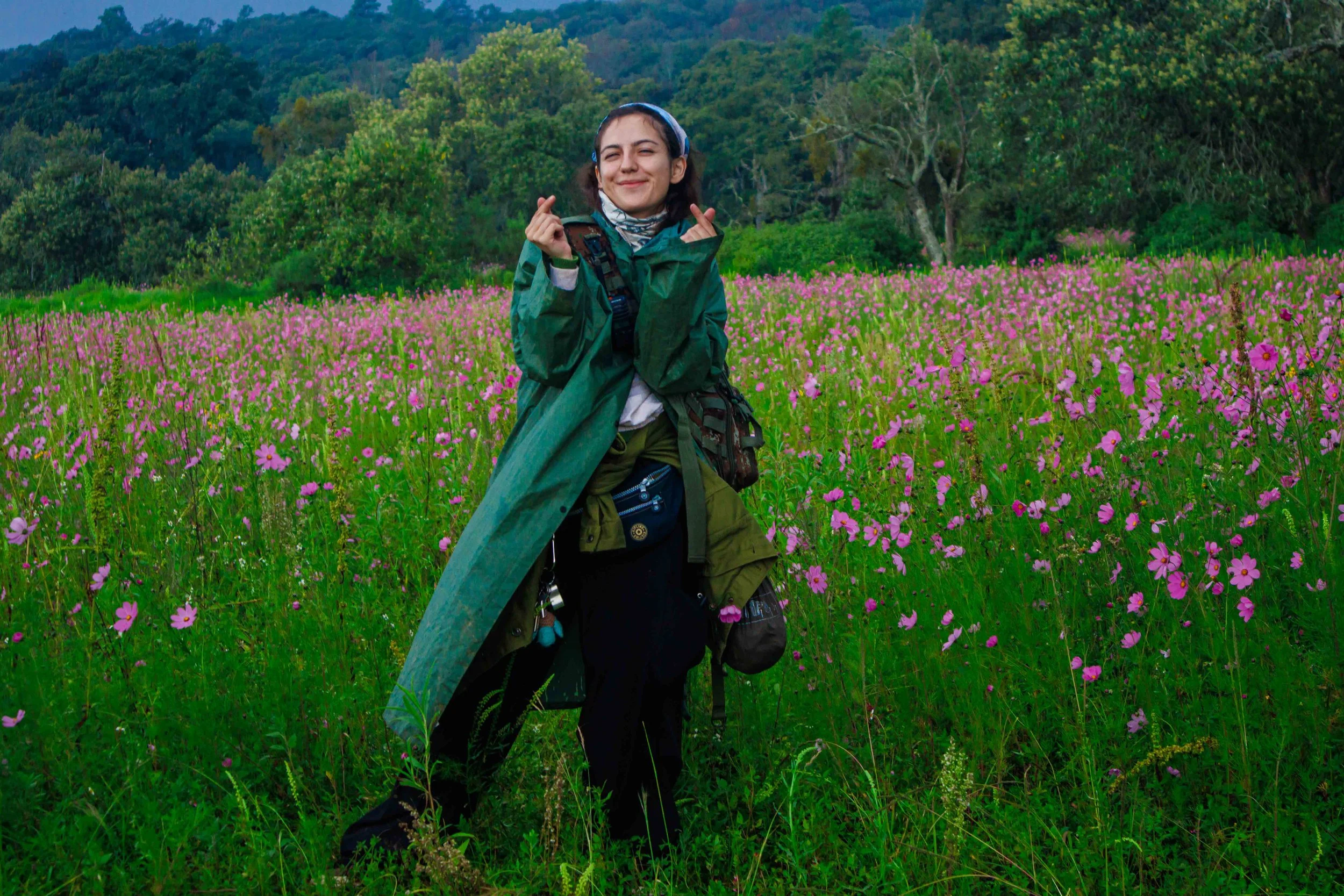 Joven sonriente en campo de flores pink, con ropa de senderismo y mochila, en medio de naturaleza verde y árboles.