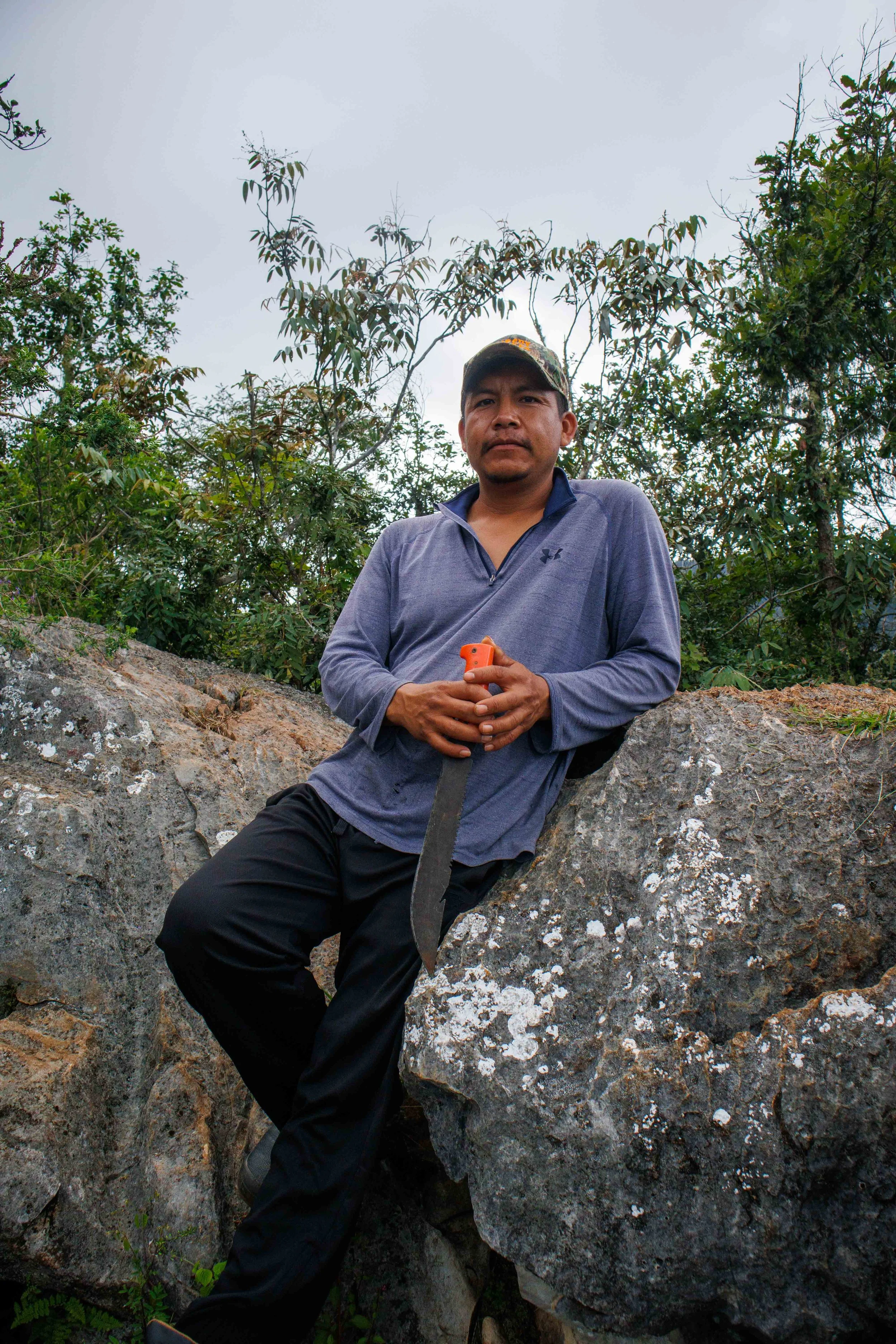 Hombre de camisa gris oscuro con gorra de camuflaje y pantalones negros está sentado sobre rocas en un área natural con árboles y vegetación al fondo, sosteniendo un objeto de color naranja en las manos.