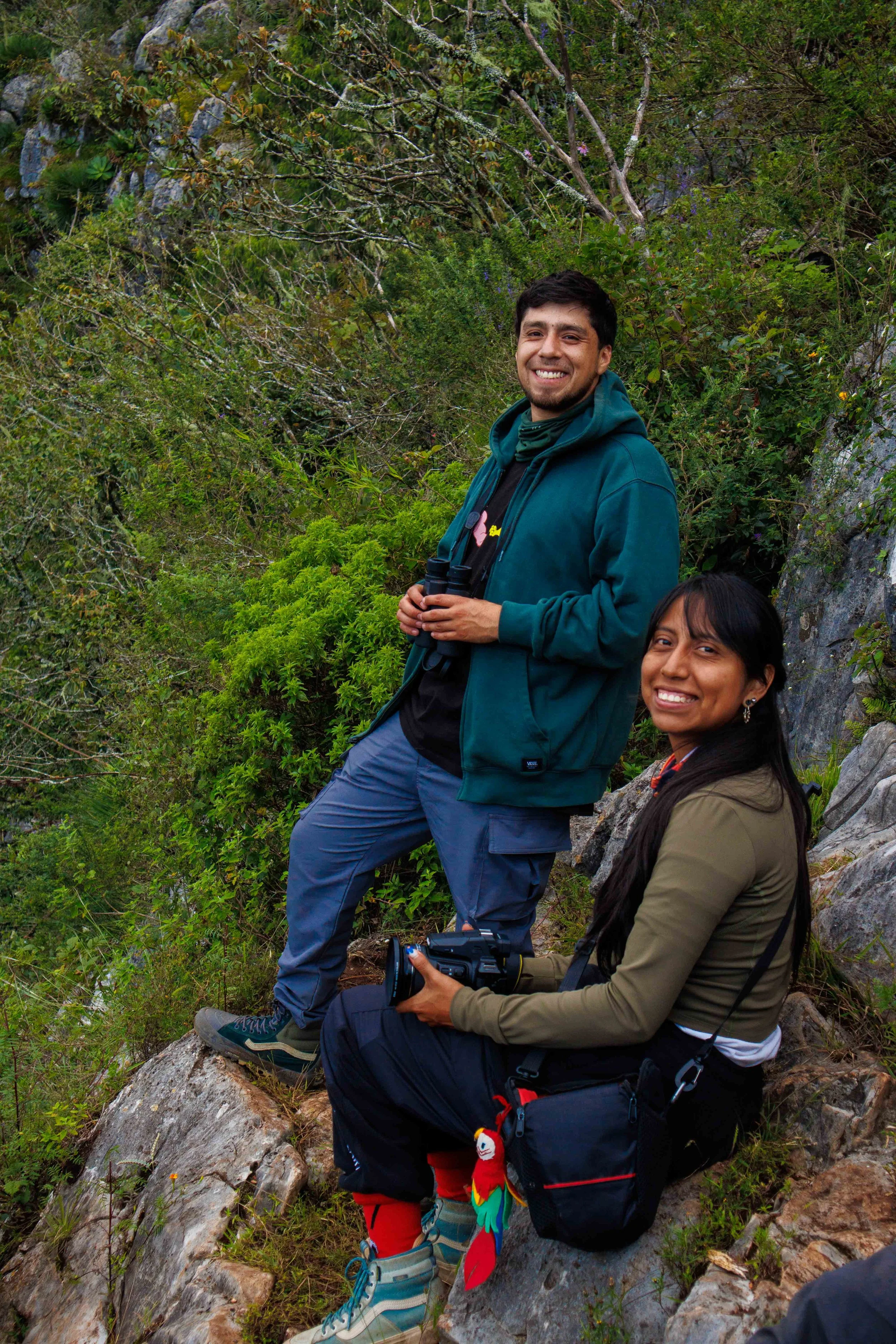 Dos personas sentadas en una roca en medio de un paisaje boscoso, una mujer sostiene una cámara y un hombre con binoculares en las manos, ambos sonrientes