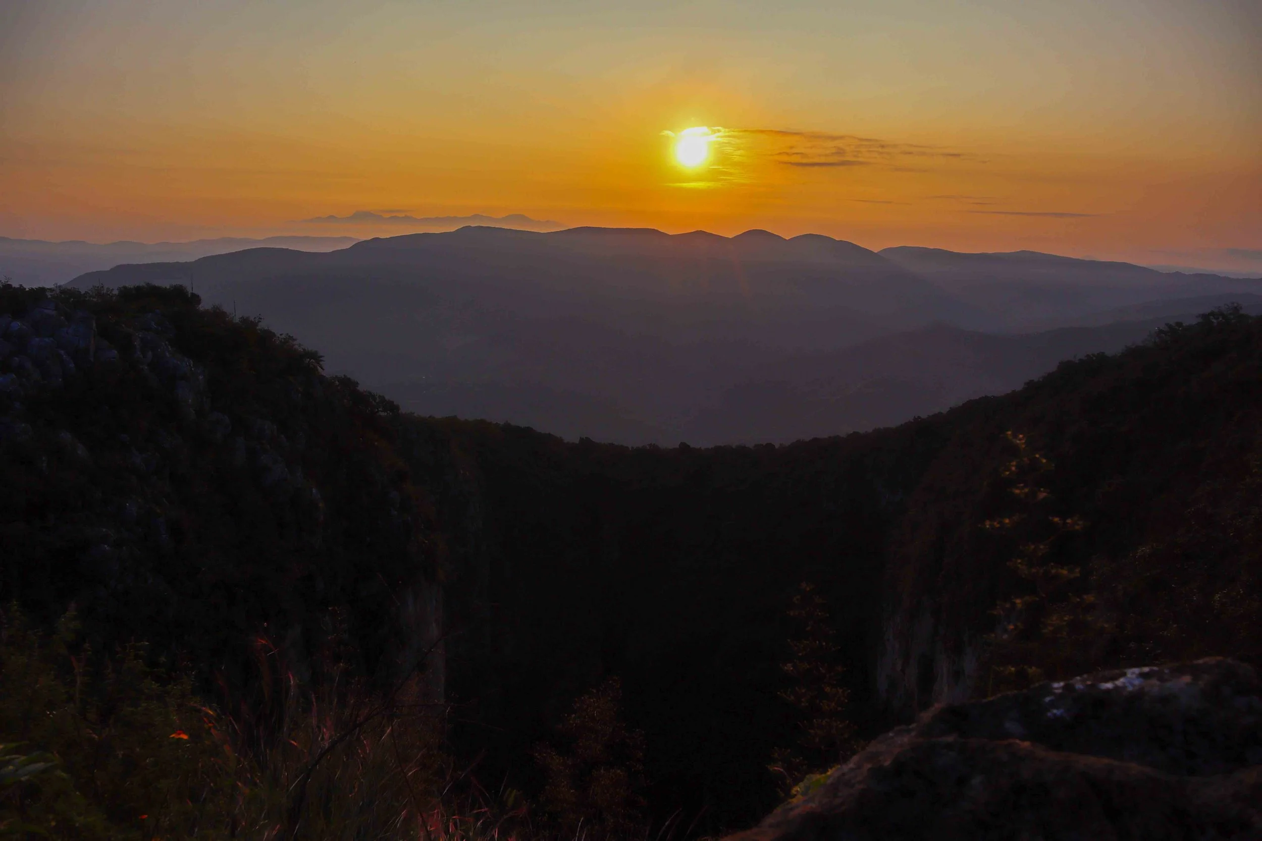 Vista de un atardecer en las montañas, con el sol parcialmente oculto por una nube y un cielo de tonos anaranjados y amarillos.