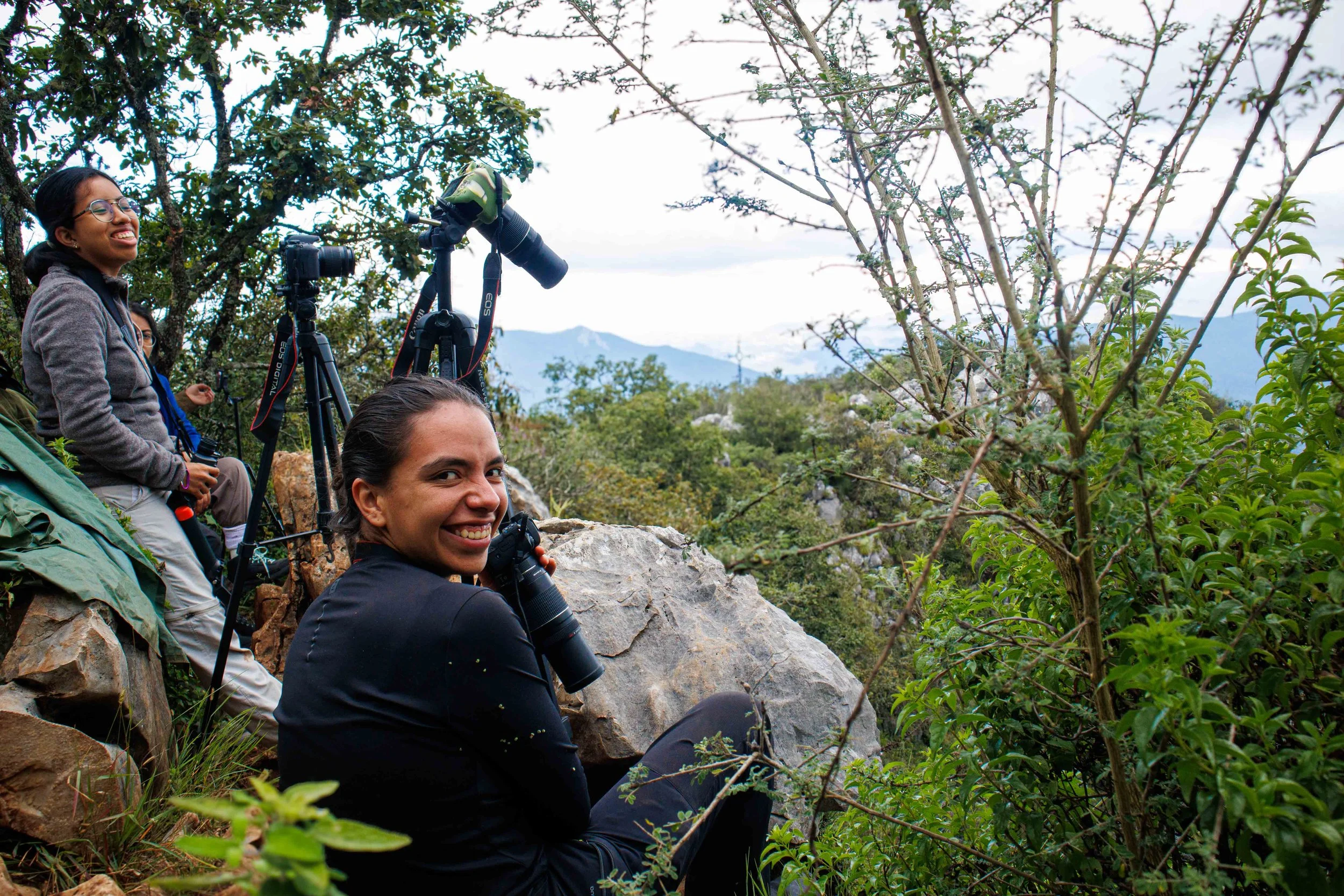 Grupo de turistas en una excursión en la naturaleza, algunos con cámaras y binoculares, rodeados de árboles y rocas en un paisaje montañoso.