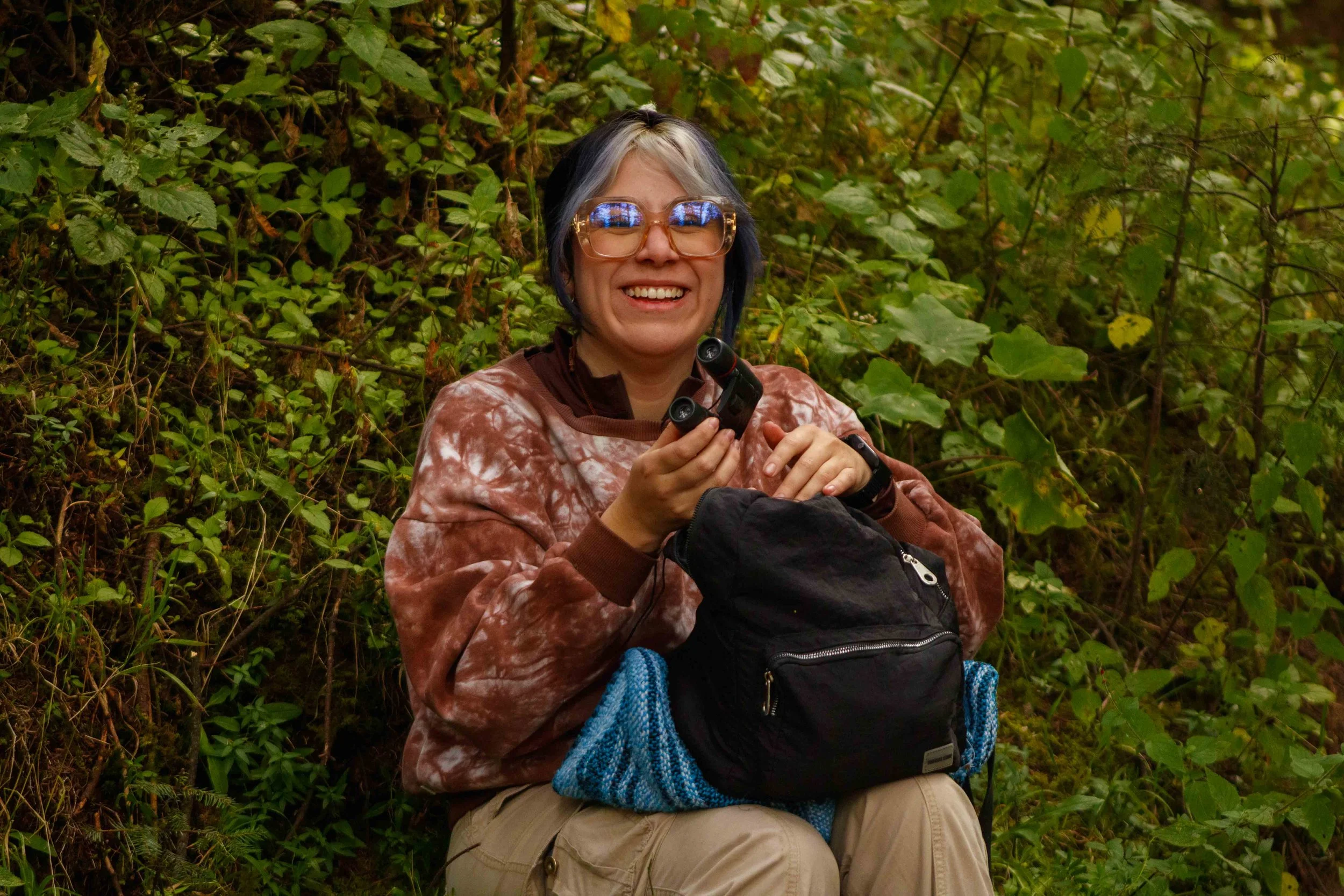 Una mujer sonriendo en un entorno de vegetación, sosteniendo binoculares y con una mochila