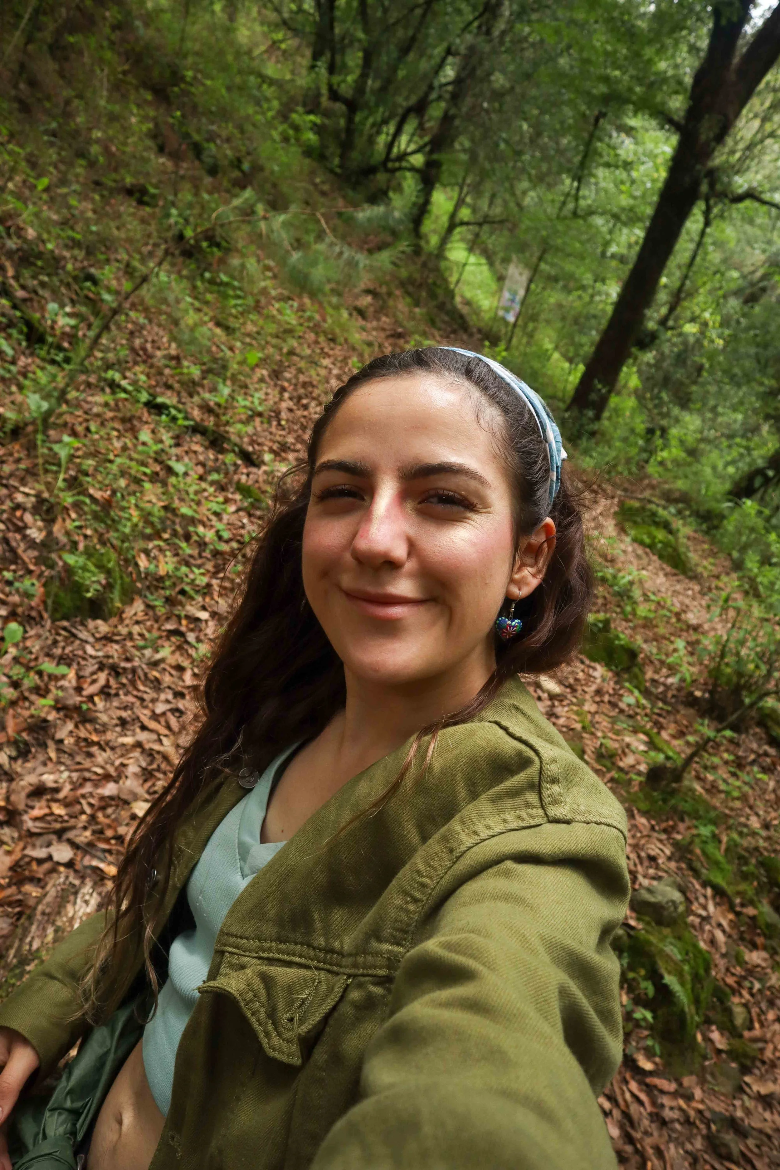 Retrato de una mujer sonriendo en un bosque con árboles y hojas caídas en el suelo.