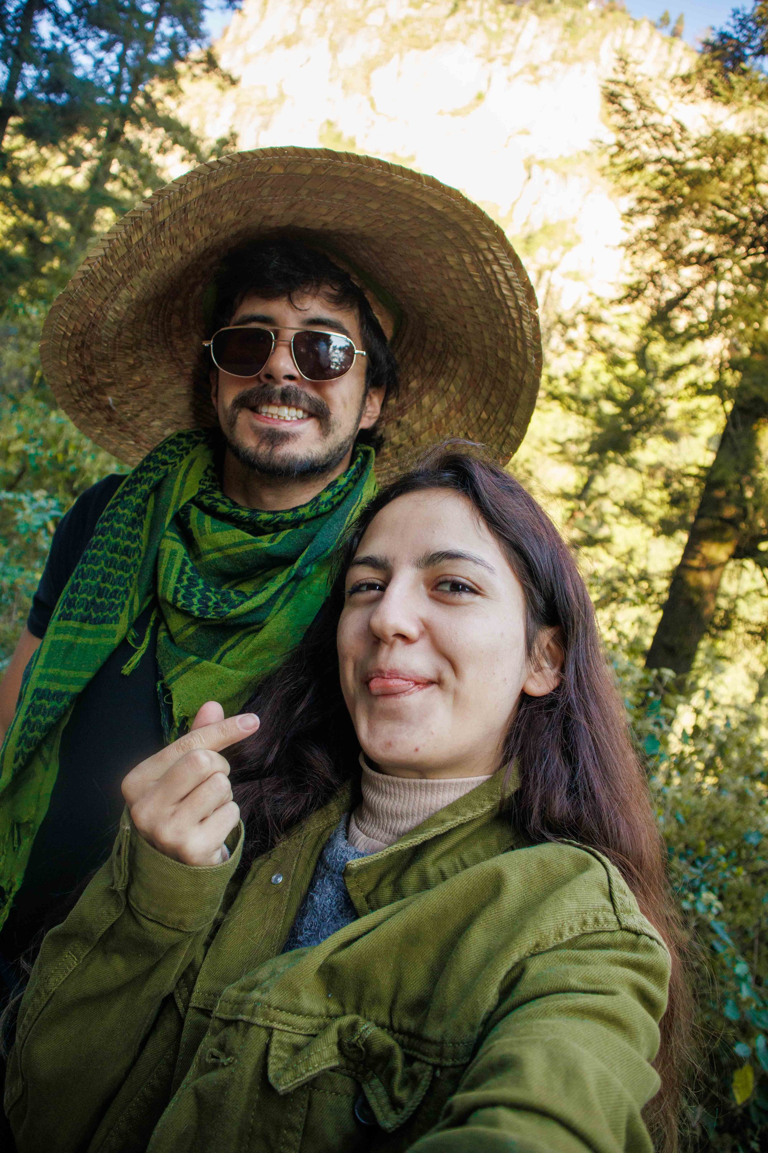 Dos personas tomando una selfie al aire libre en un entorno natural con árboles y colinas en el fondo, una mujer en primer plano sonriendo y haciendo una señal de paz, un hombre con sombrero amplio, gafas de sol y bufanda sonriendo al fondo.