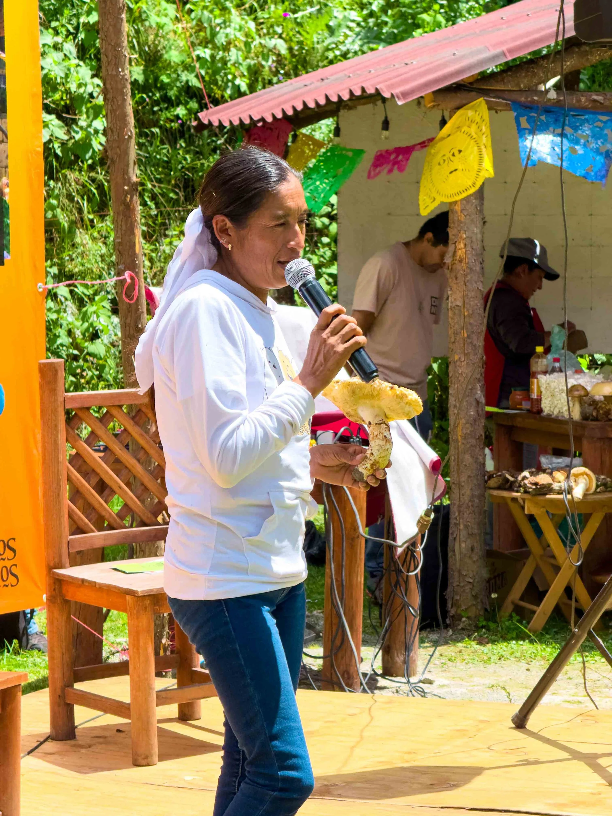 Mujer con blusa blanca y pantalones de mezclilla sosteniendo un micrófono en un ambiente al aire libre con decoración colorida y dos personas en el fondo trabajando en un puesto de comida.