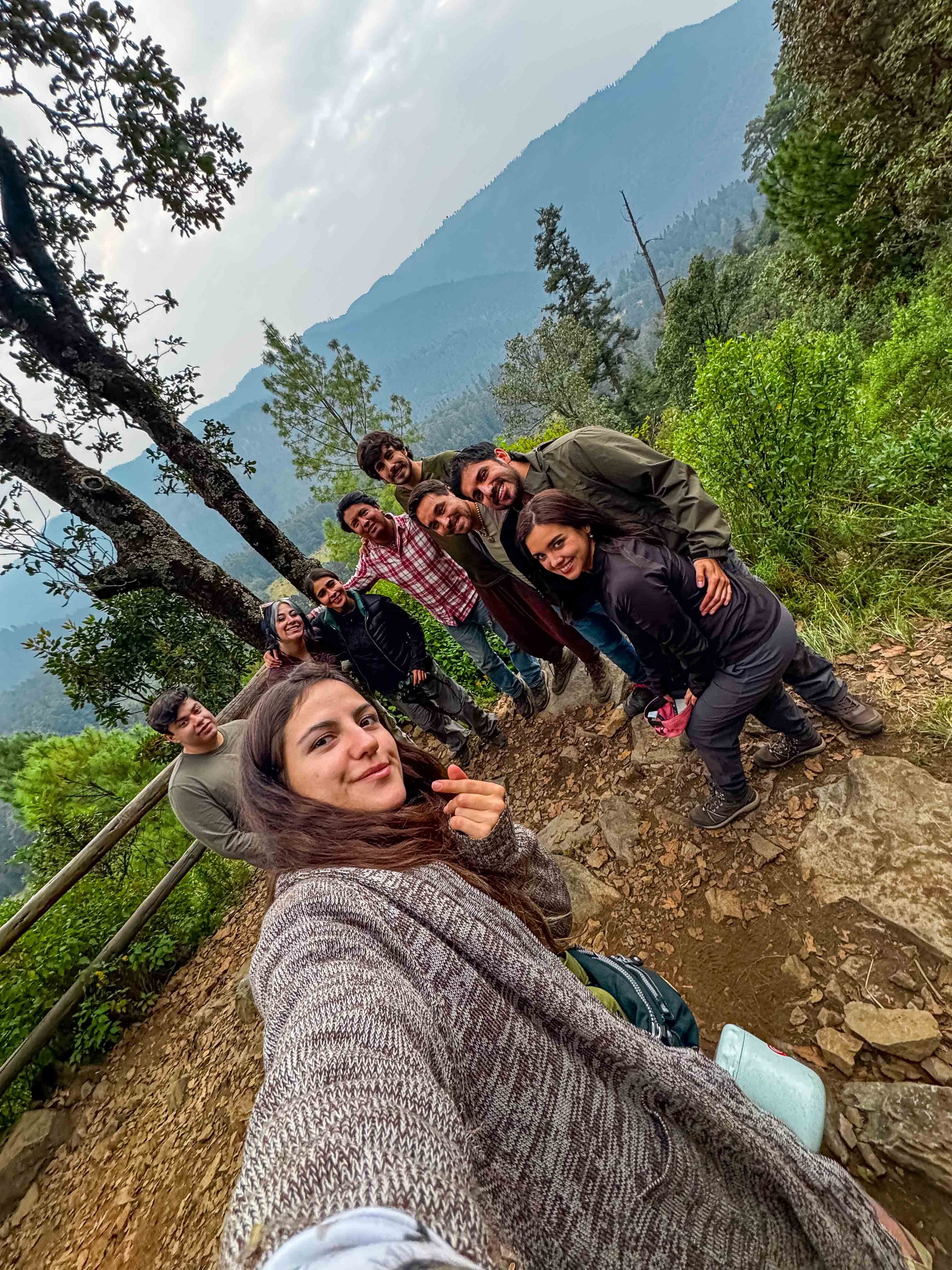 Un grupo de personas en un sendero de naturaleza con árboles y montañas de fondo, sonriendo y posando para una selfie.
