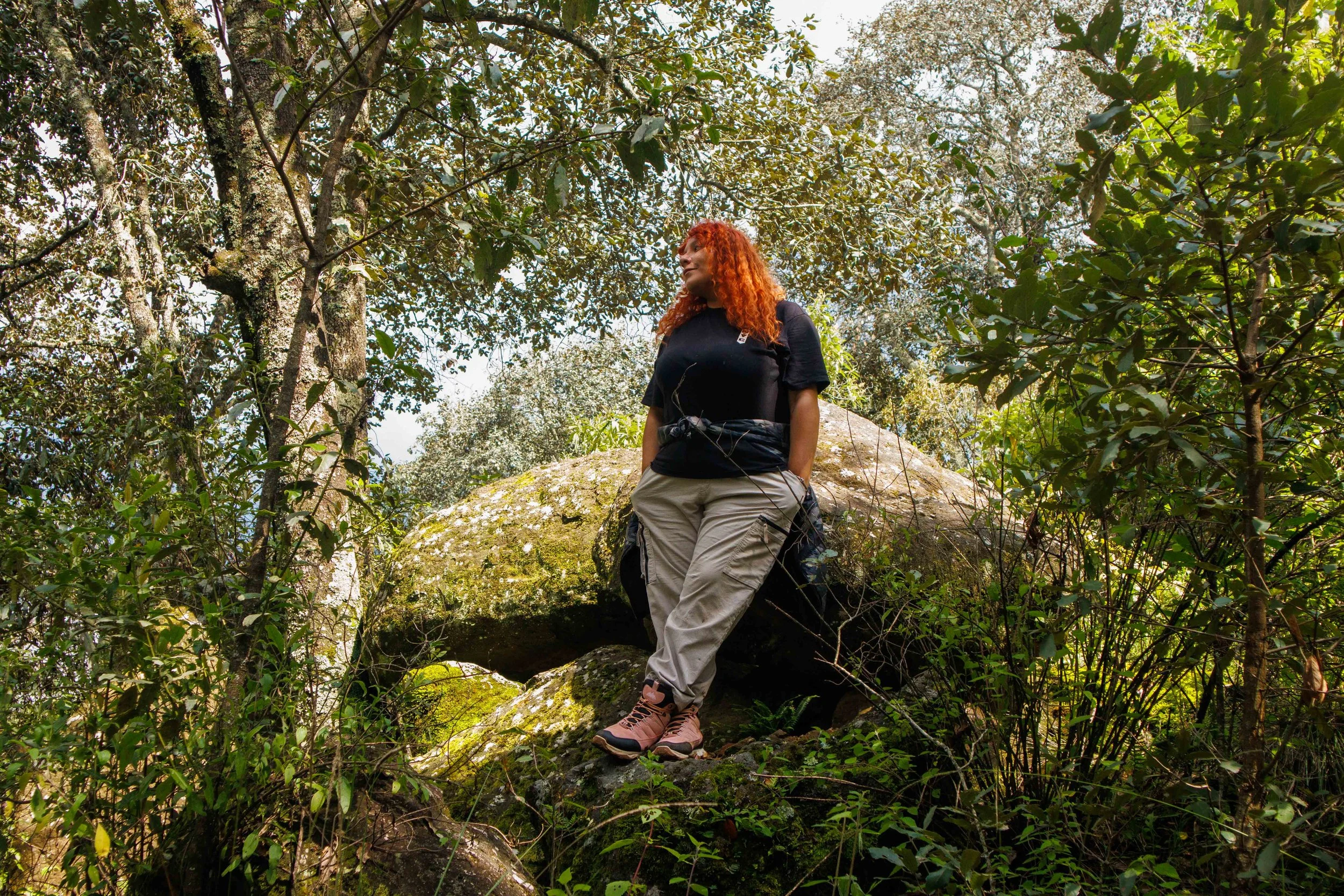 Una mujer con pelo rizado y rojo, vistiendo ropa de senderismo, de pie sobre una piedra en un bosque con árboles verdes.