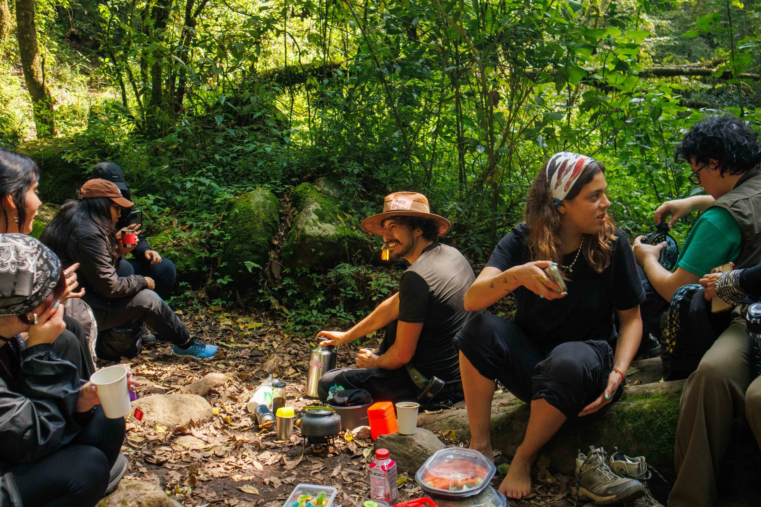 Grupo de personas sentadas en el suelo de un bosque, conversando y compartiendo comida y bebida, rodeados de vegetación espesa y árboles.