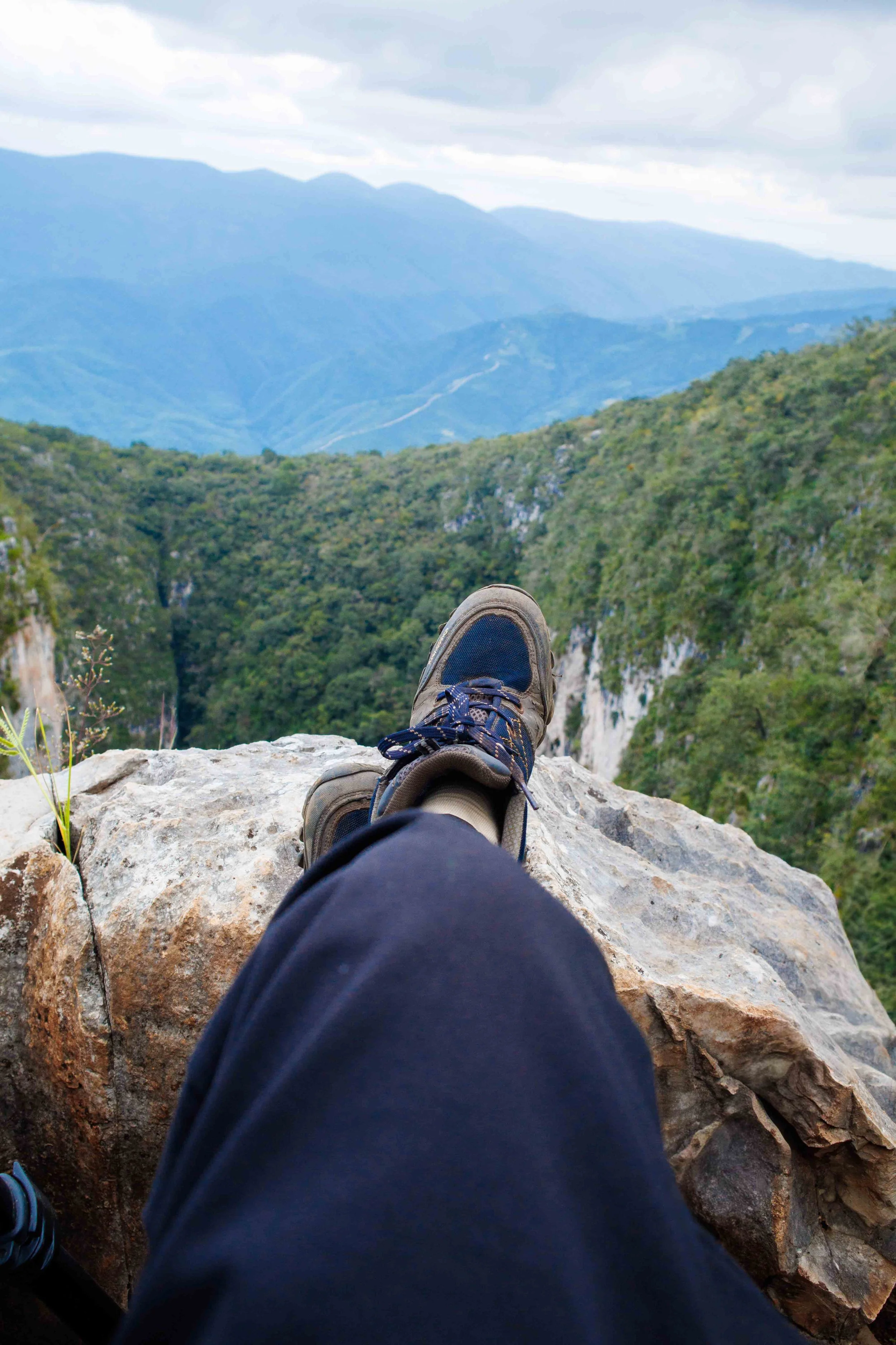 Persona acostada sobre una roca, con vista a un valle y montañas al fondo, usando zapatos de senderismo y pantalones oscuros.