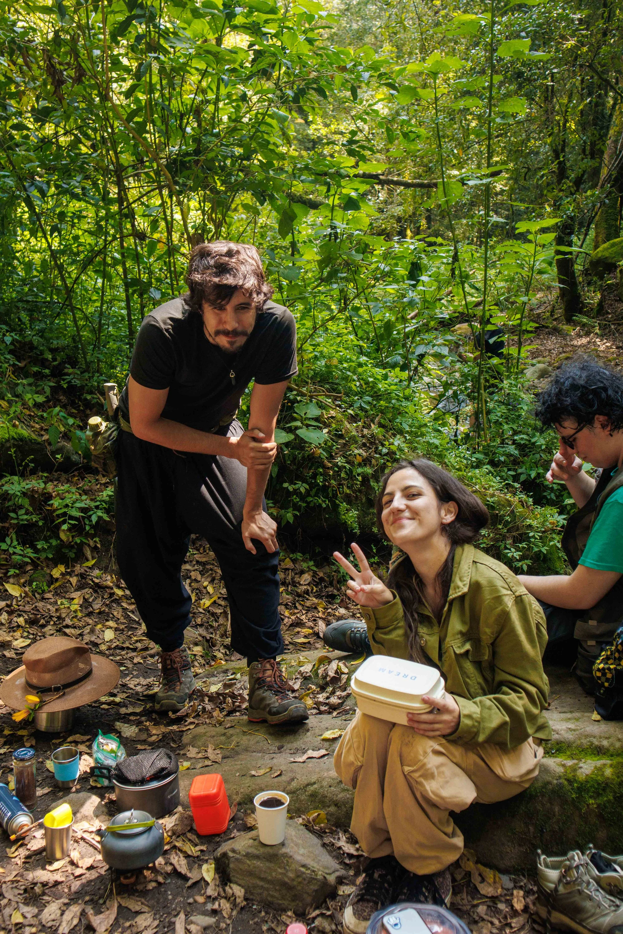 Un grupo de personas disfrutando de un día en la naturaleza en un bosque, con varios objetos de comida y bebidas en el suelo, posando y sonriendo.