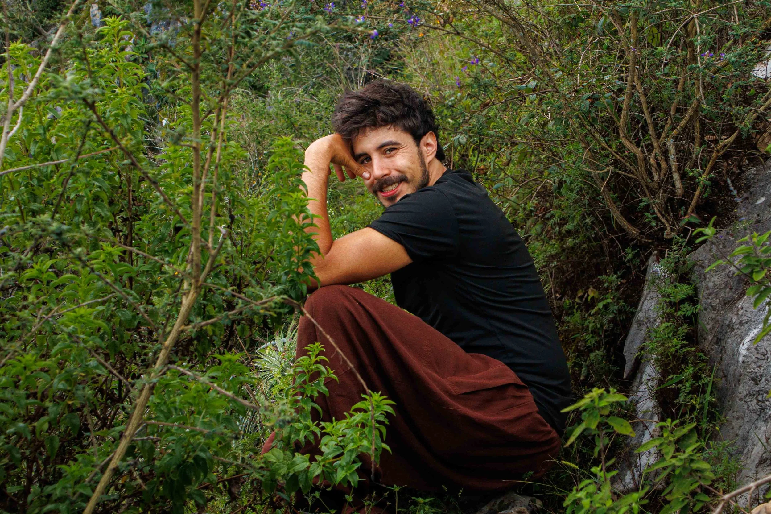Jose Luis Fernandez Coello (Monte) de AlMonte Hike . Joven sentado en bosque rodeado de vegetación verde y arbustos, con expresión sonriente, mirando hacia la cámara.