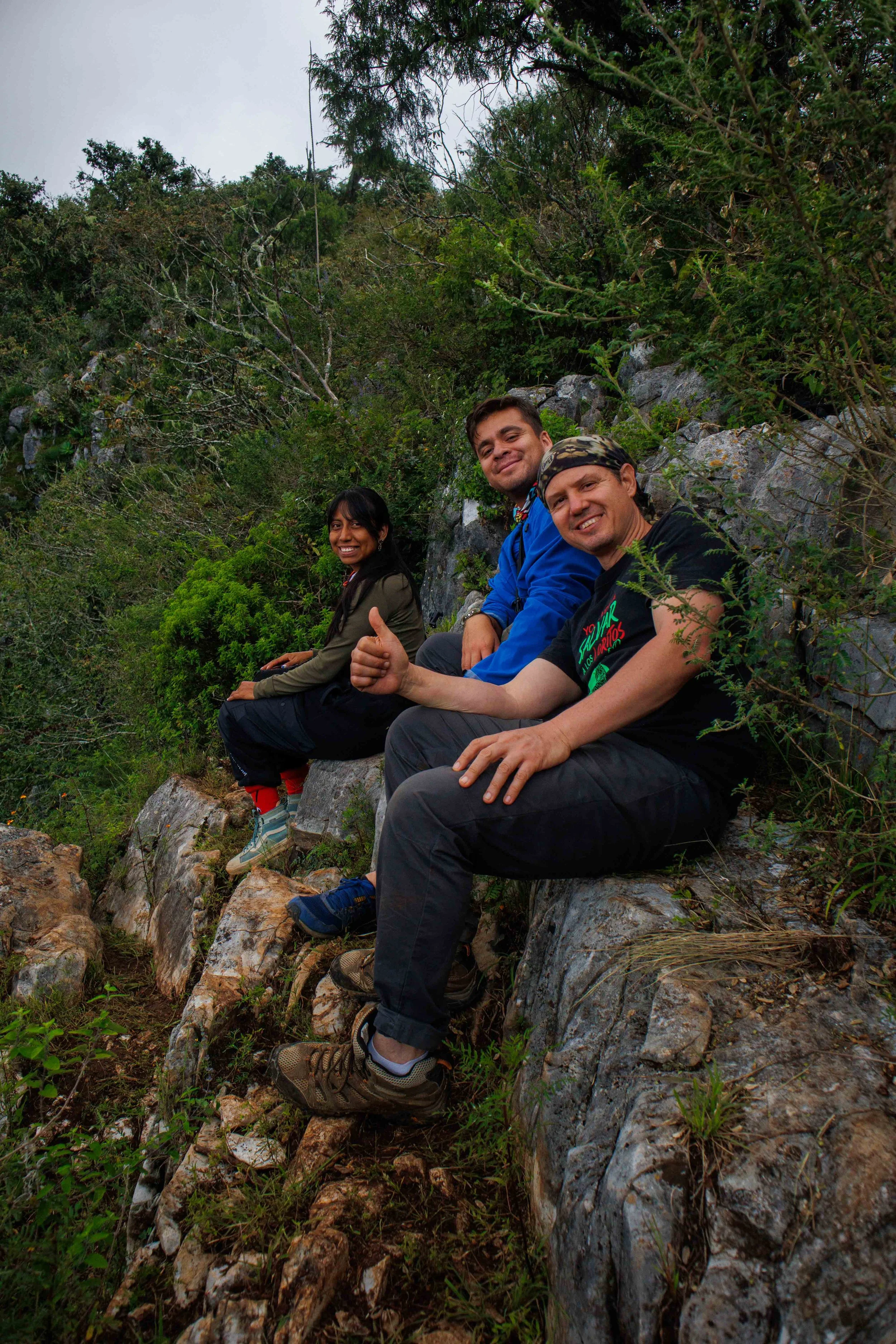 Tres personas sentadas sobre rocas en un entorno natural con vegetación densa y árboles durante el día, una de ellas dando un pulgar hacia arriba y sonriendo esperando para fotografiar guacamayas en la reserva de la biosfera de la Sierra Gorda.